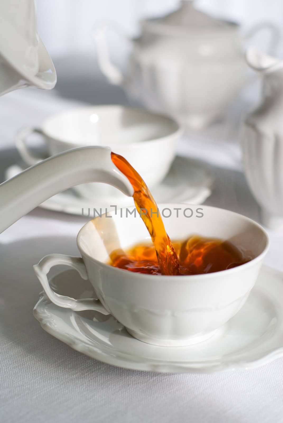 Pouring fresh tea from porcelain tea kettle into white cup. Focus on tea stream, shallow depth of field.