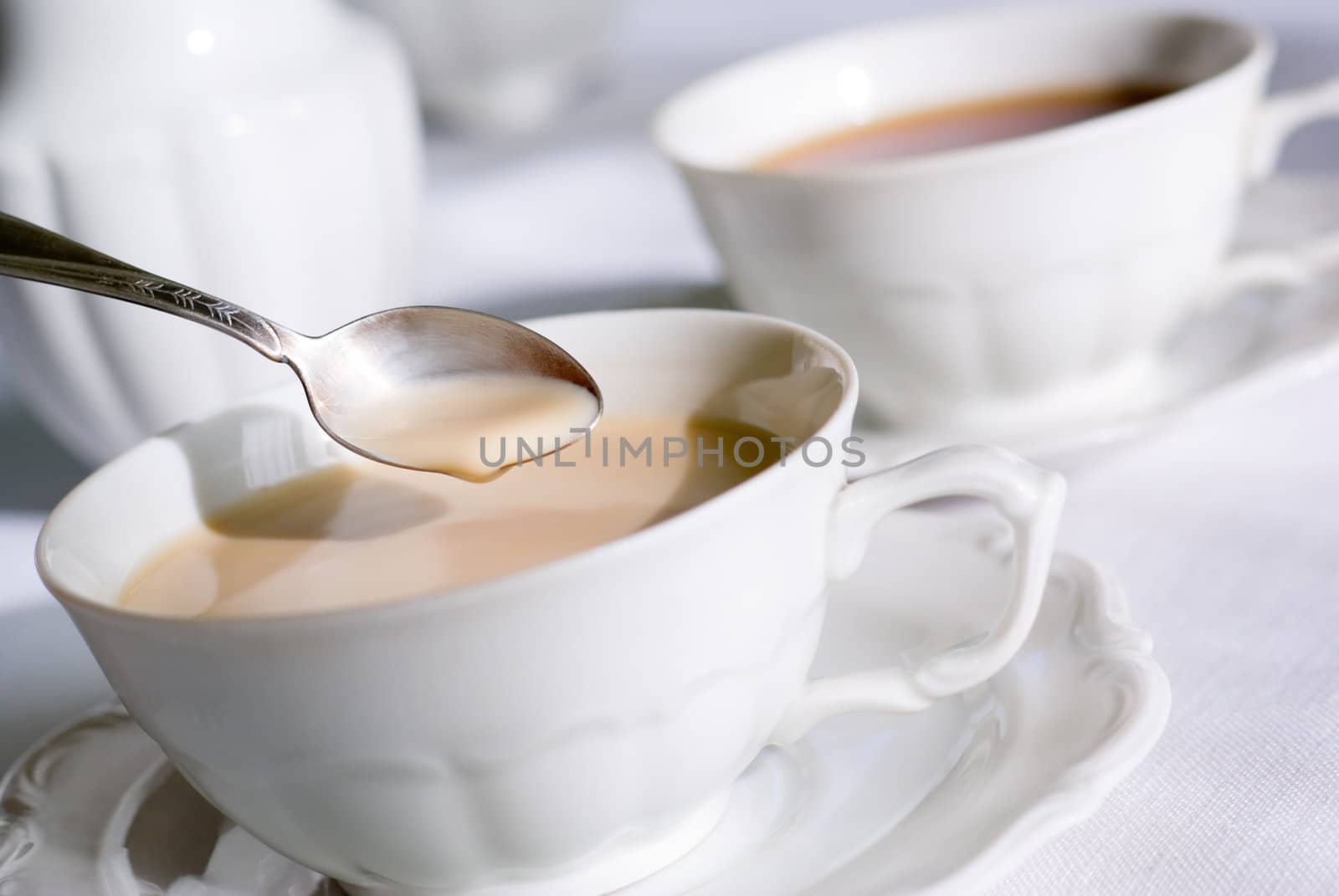 Tea or coffee set - teaspoon over two porcelain cup on the table, one with milk. Shallow depth of field (focus on teaspoon).