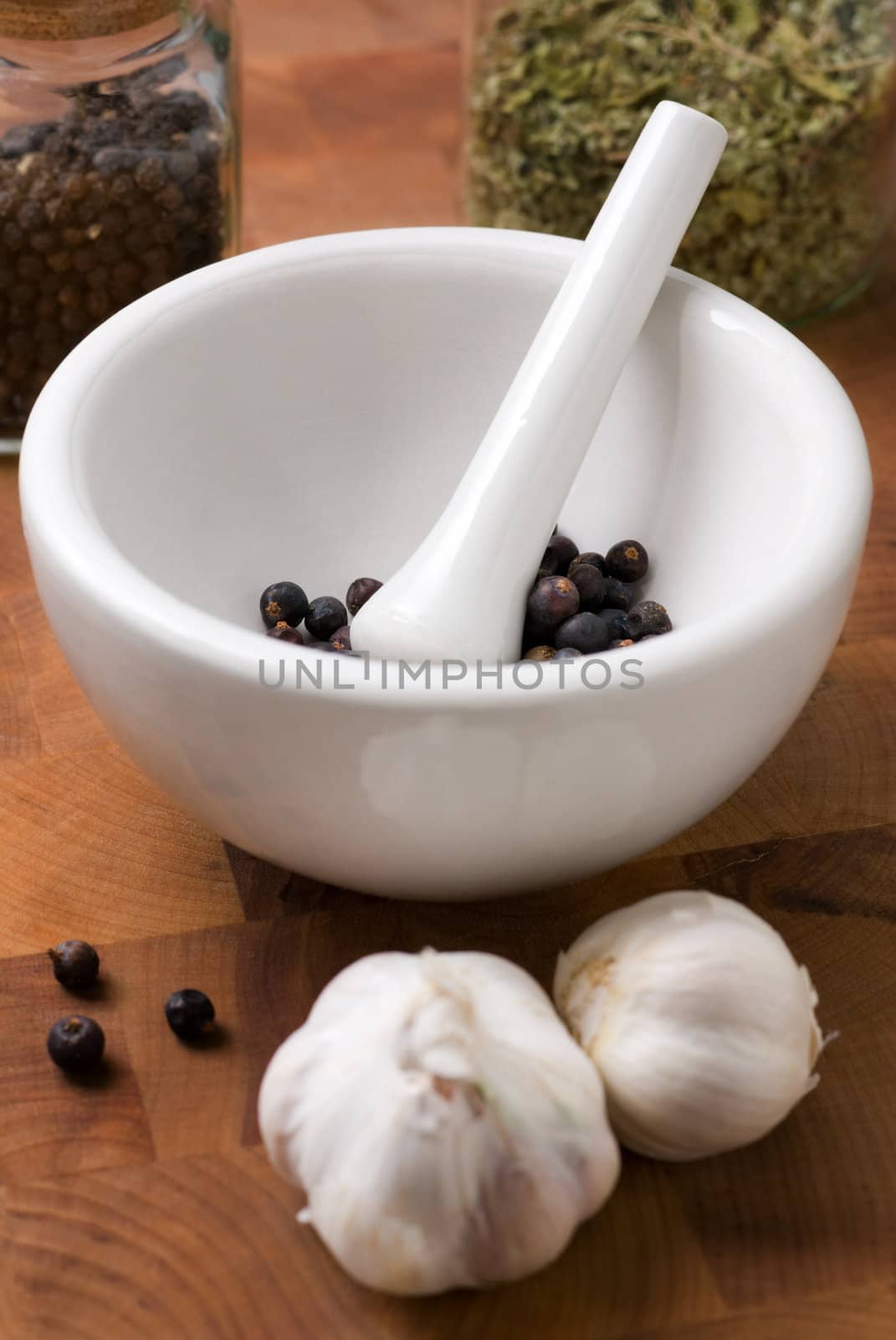 Juniper (focus on it) in ceramic mortar and pestle and garlic on the wood cutting board. Two jars with herb and spice behind.