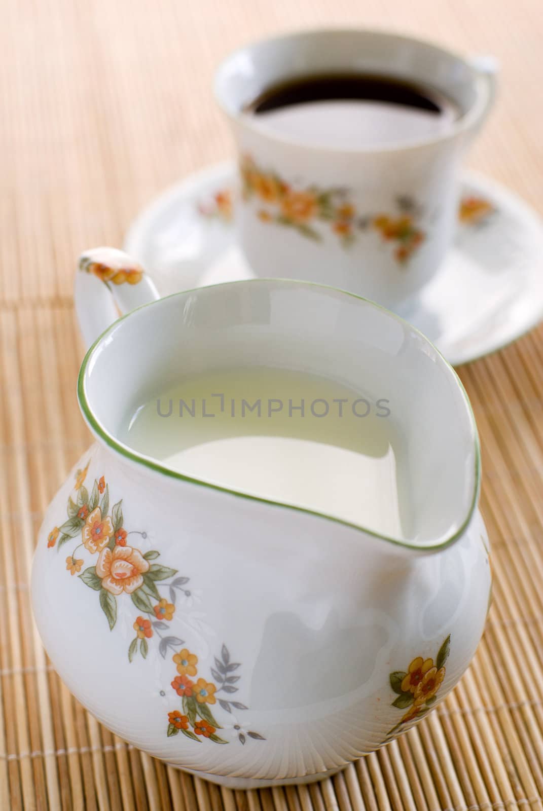 Milk in floral pattern porcelain milk jug on the wooden mat - on the front and cup of coffee behind. Shallow DOF.