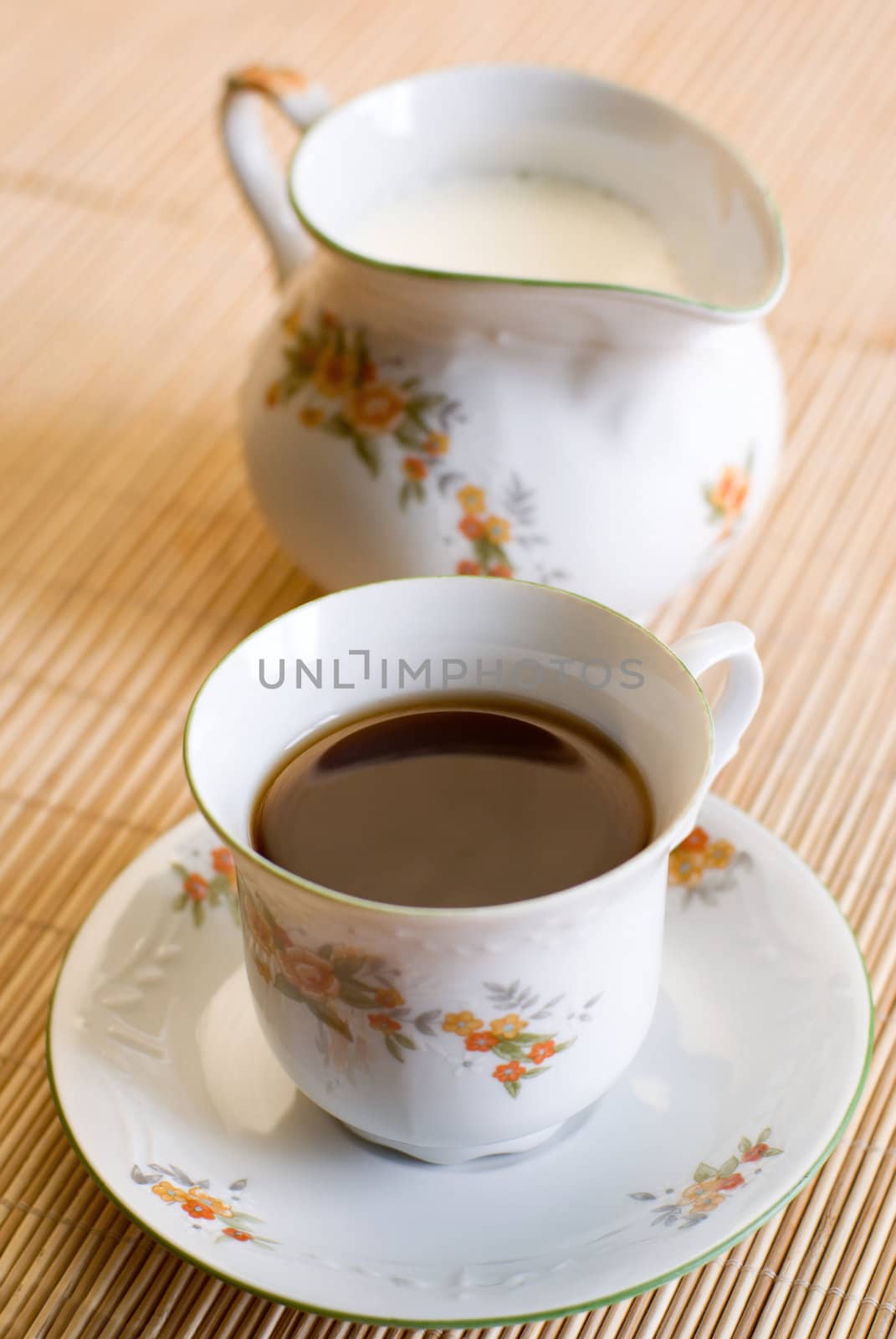 Cup of coffee or black tea (on the front, focus on the liquid surface) and a floral pattern porcelain milk jug on the wooden mat behind. Shallow DOF.