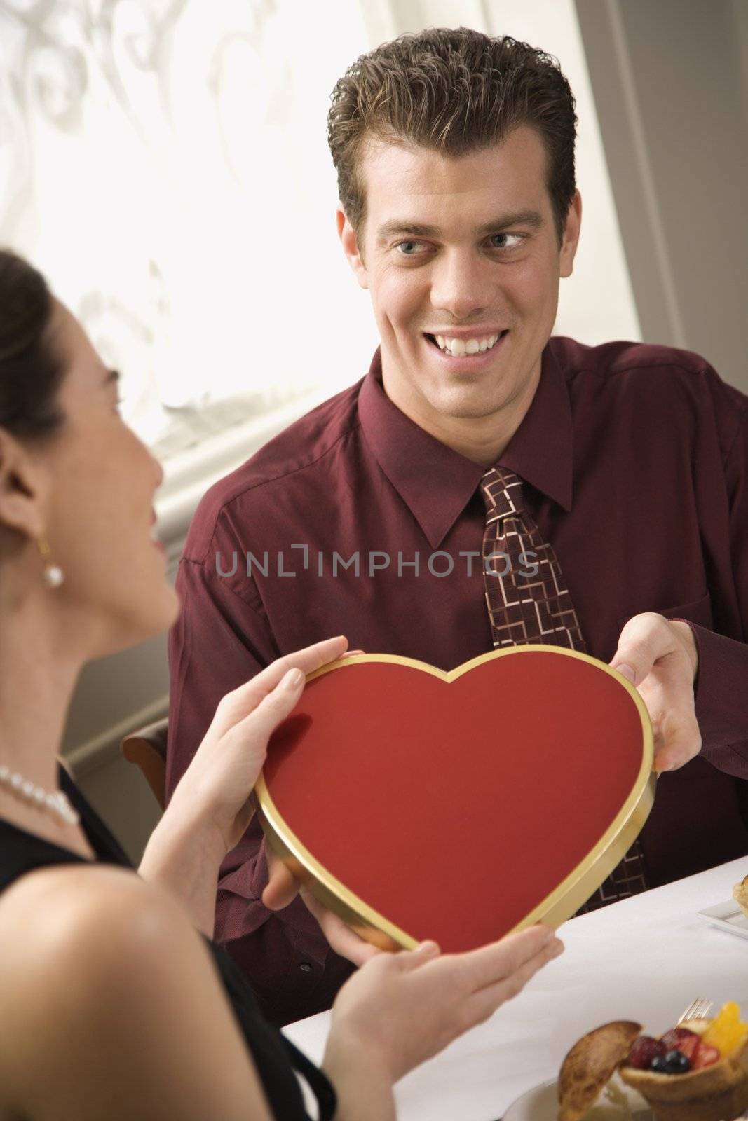 Mid adult Caucasian man giving a heart shaped box of chocolates to surprised woman at restaurant.