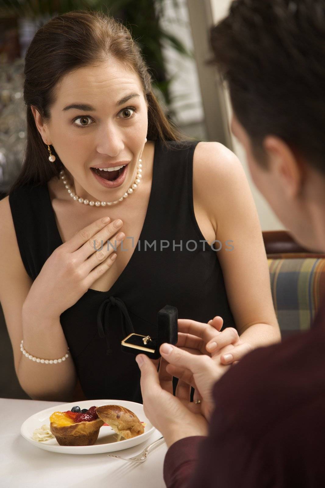 Mid adult Caucasian man proposing marriage to surprised woman at a restaurant.