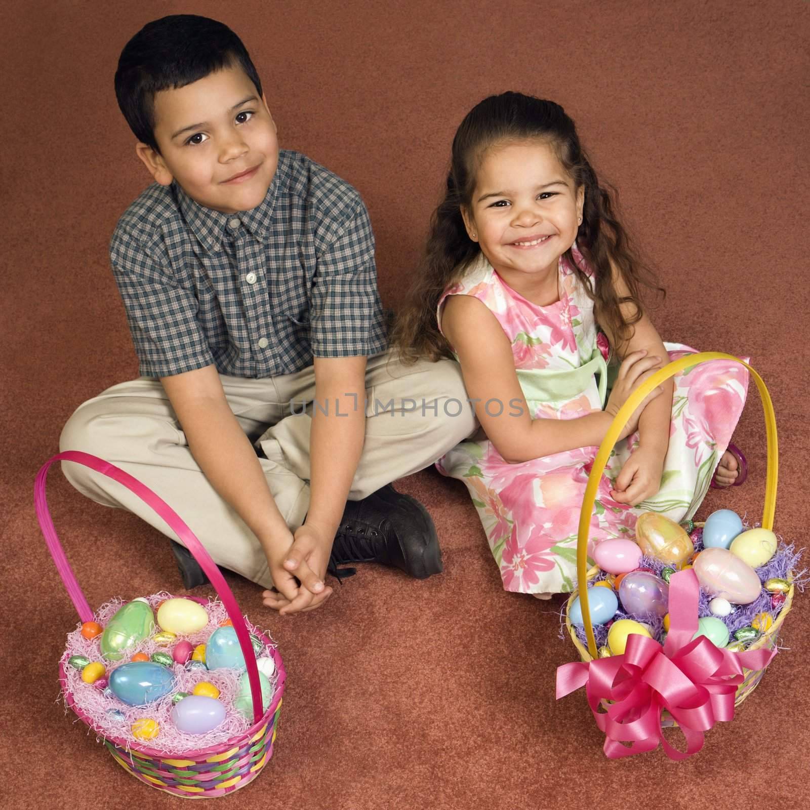 Hispanic brother and sister sitting on floor with Easter baskets looking up at viewer smiling.