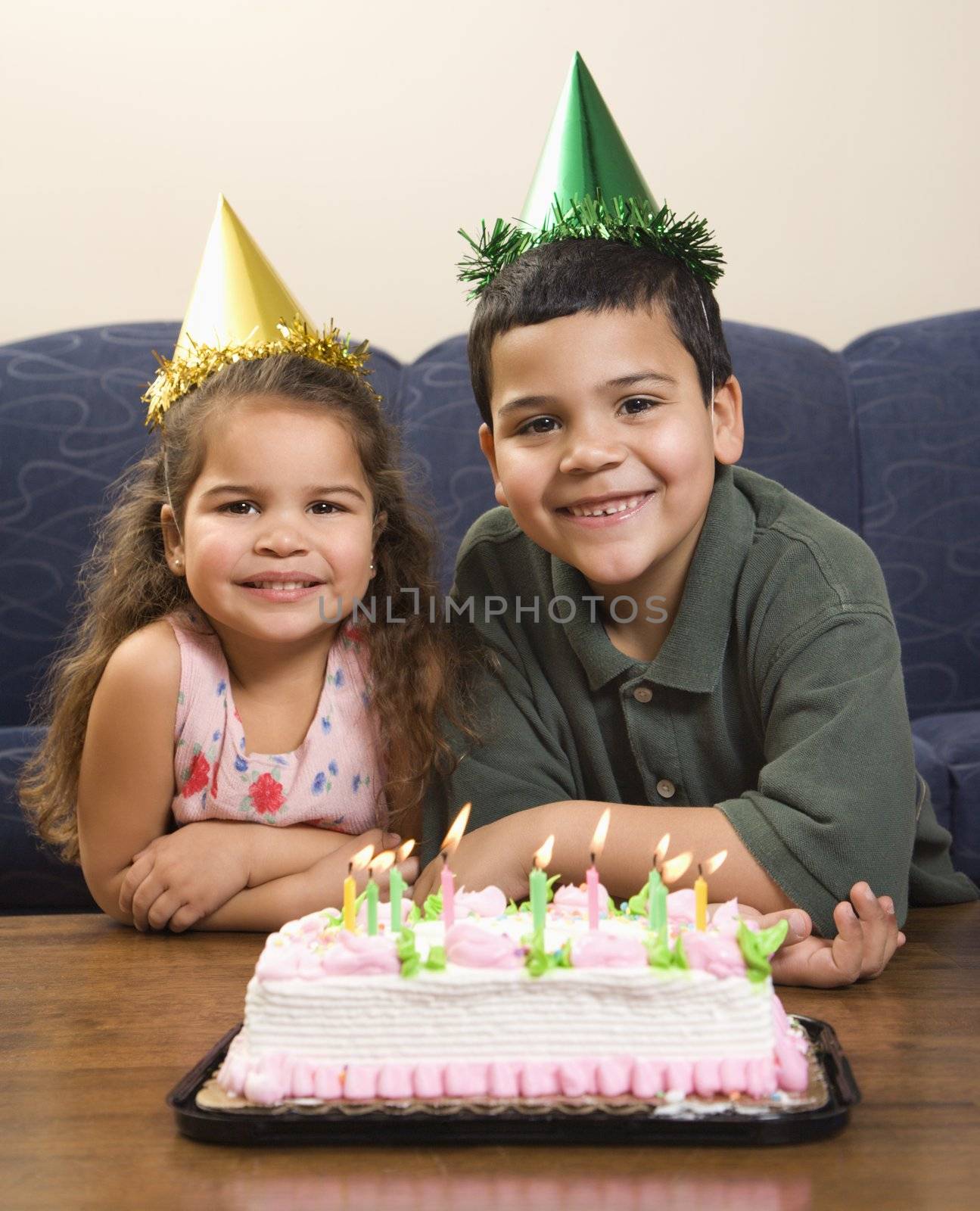 Hispanic girl and boy wearing party hats sitting in front of birthday cake smiling and looking at viewer.