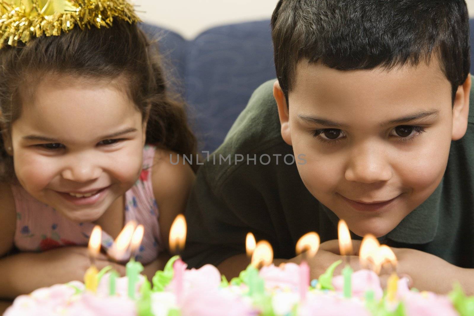 Hispanic girl and boy leaning in close looking at lit candles on birthday cake and smiling.