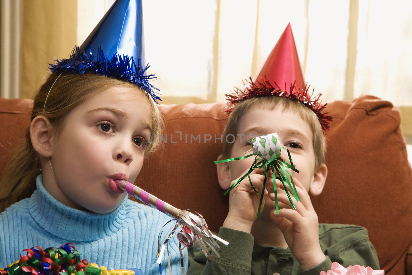 Caucasian children looking bored wearing party hats and blowing noisemakers.