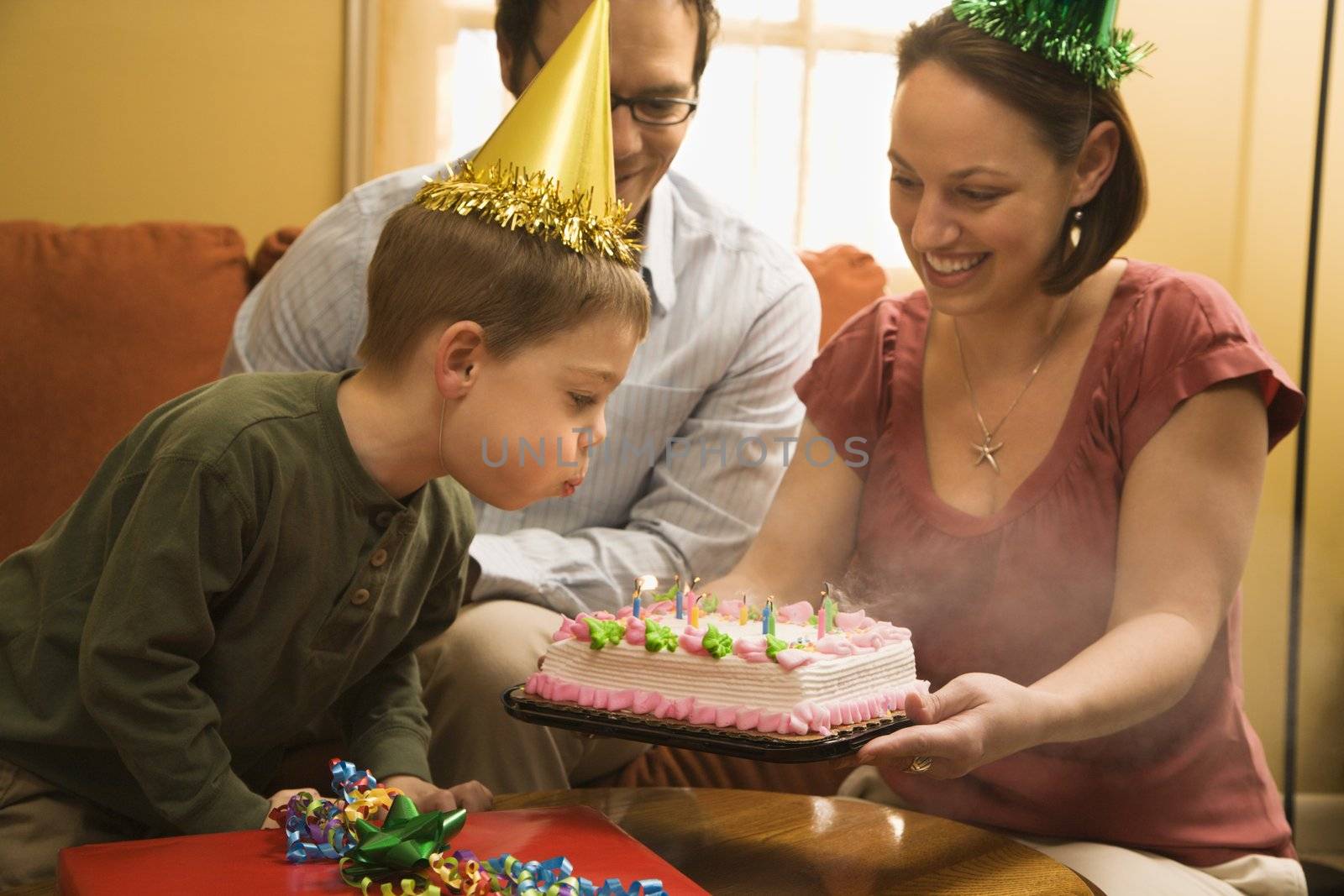 Caucasian boy in party hat blowing out candles on birthday cake with family watching.