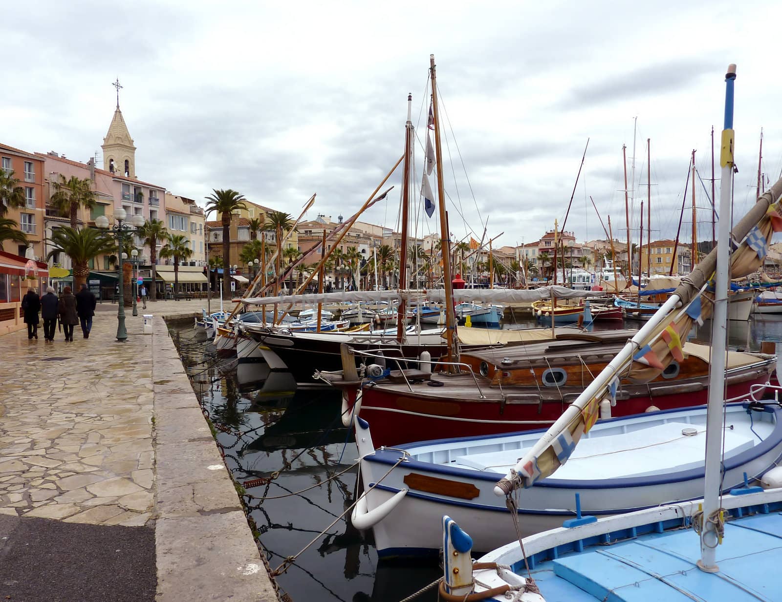 View of Sanary-sur-mer harbor with its colored boats and people walking on the side with a church, palm trees and buildings, France, by cloudy weather