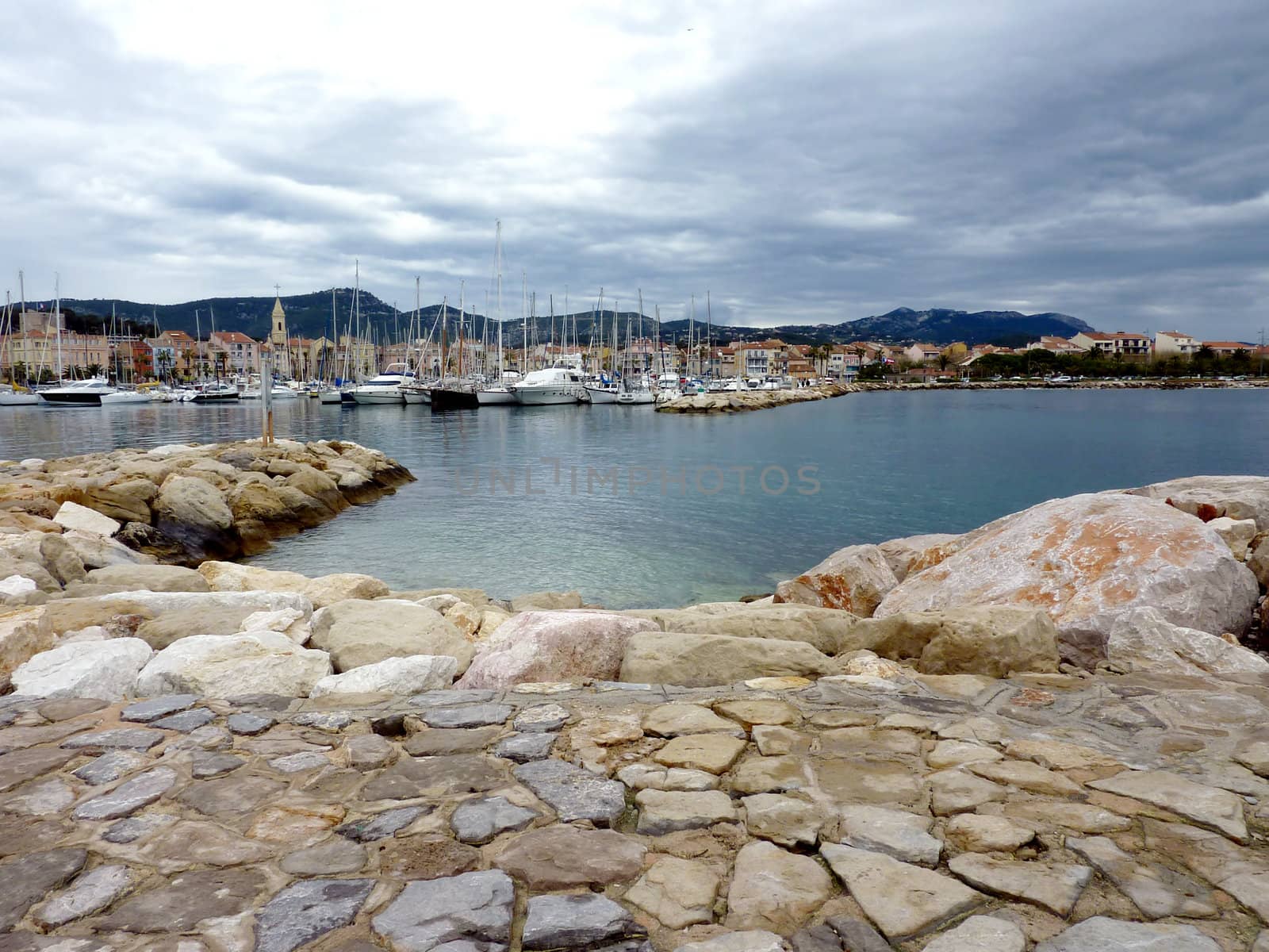 View of Sanary-sur-mer harbor with its famous church and the boats, from a place made of stones in the foreground by cloudy weather, France