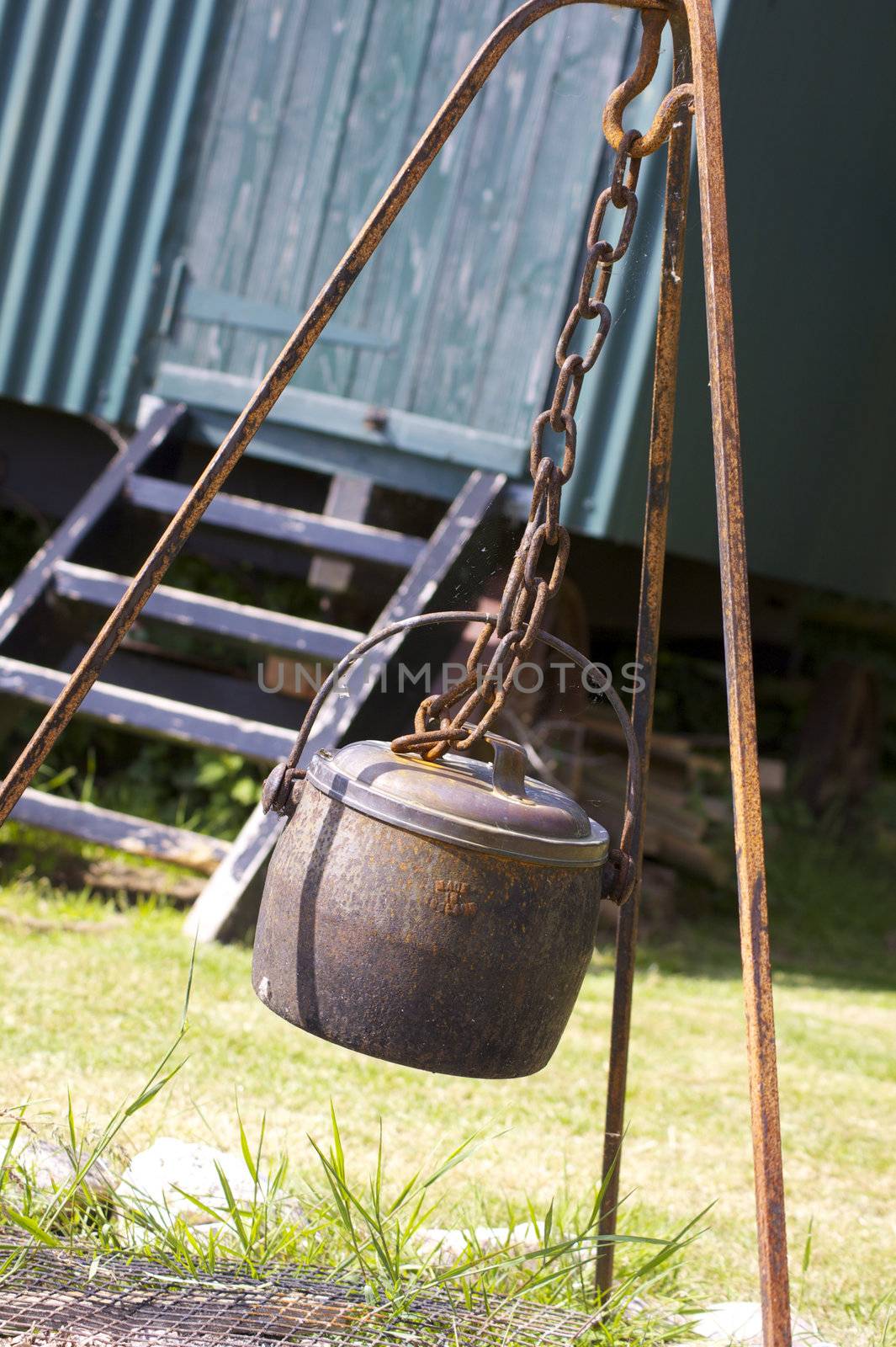 A rusty metal cauldron hanging over an open fire space within a rusty metal frame, hook and chain, set in front of the wooden steps of a green painted tin constructed shepherds hut. Located in rural Dorset, England.
