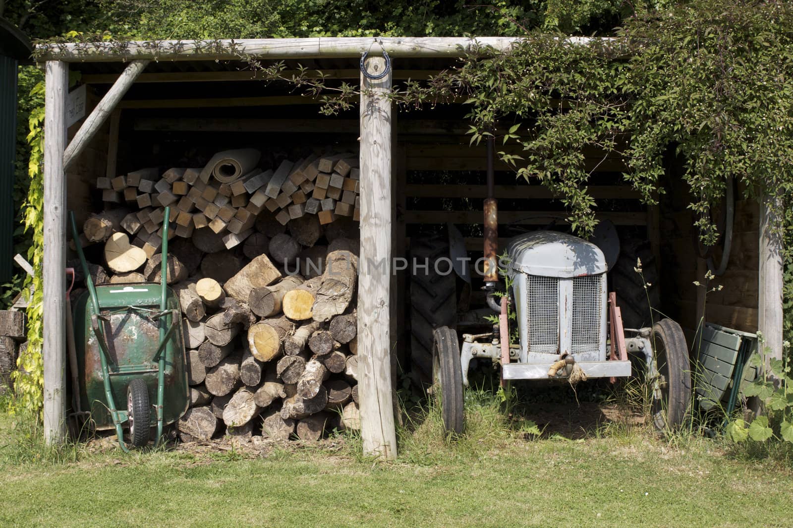 A rural farmyard wooden shed with stored cut logs to one side, with a vintage grey tractor to the other. A green wheelbarrow leant against the pile of logs.
