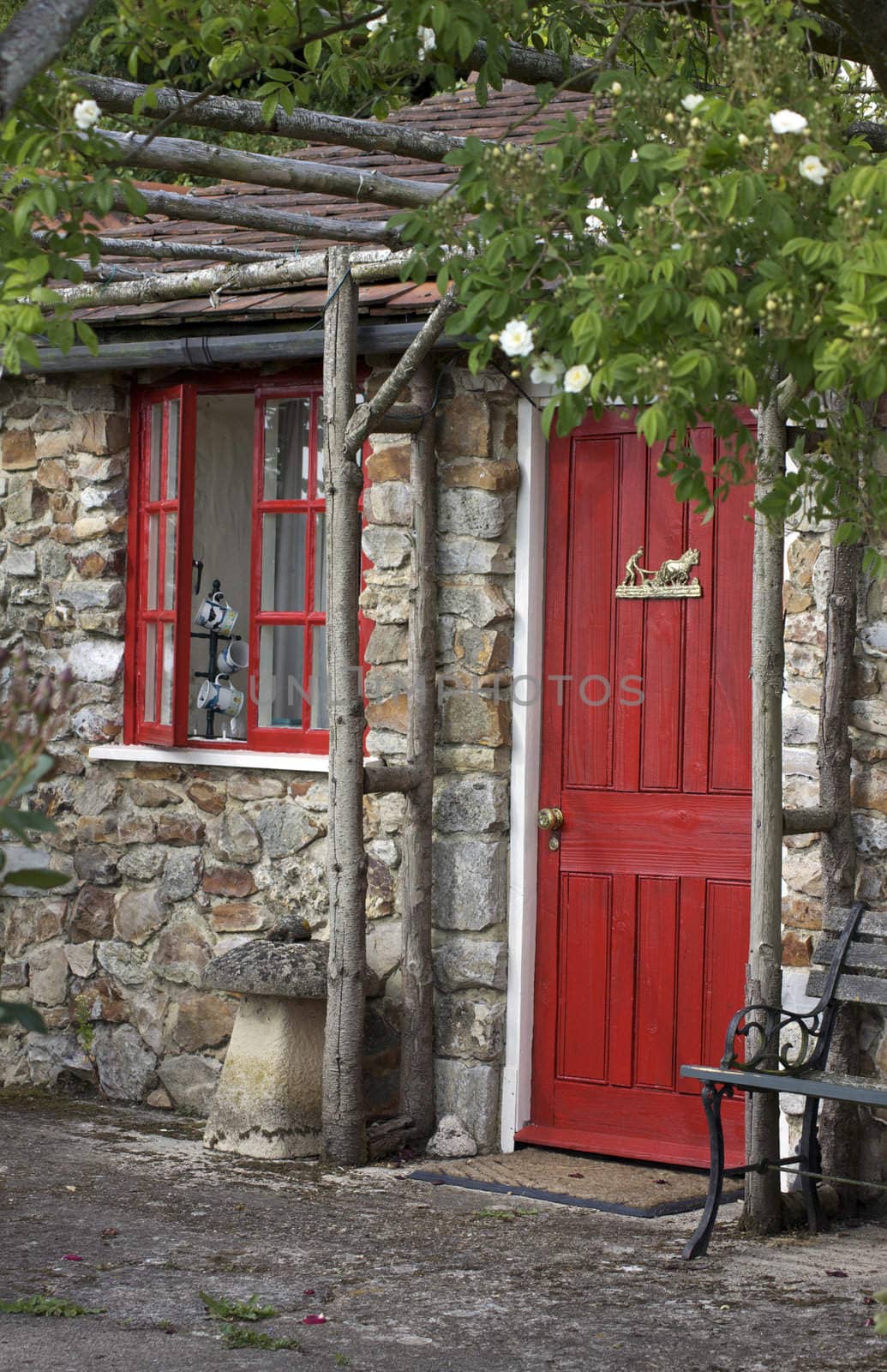 A stone constructed summer house with a terracotta slated roof, a red painted wooden door and window frames. A wooden trellice covered in white roses adorns the front. Located in Devon,England.