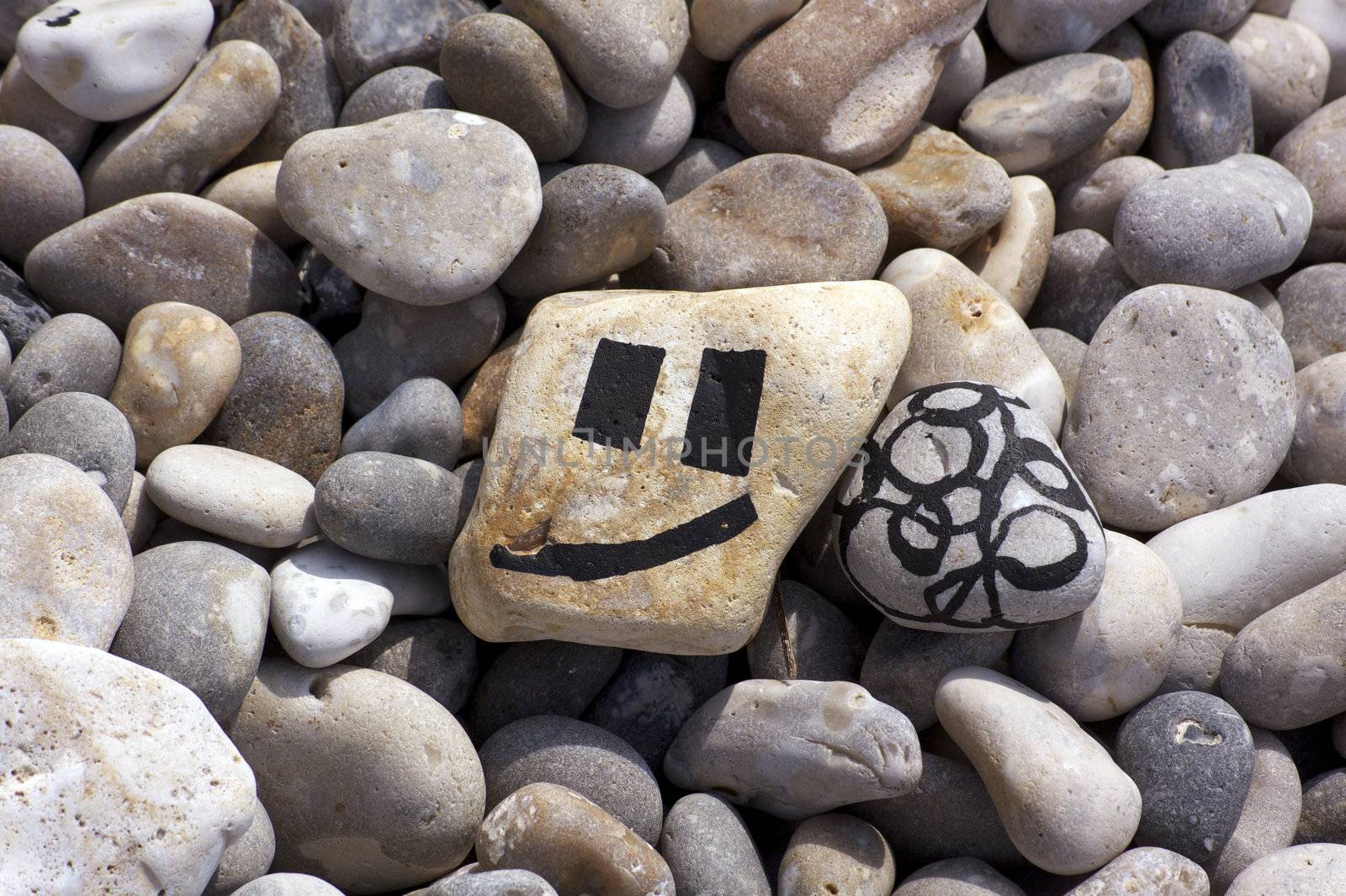Two pebbles found on the beach at Beer in Dorset, England. Both pebbles are hand painted. one with a smiley face, the other with an abstract pattern. Both lie amongst other pebbles on the beach.