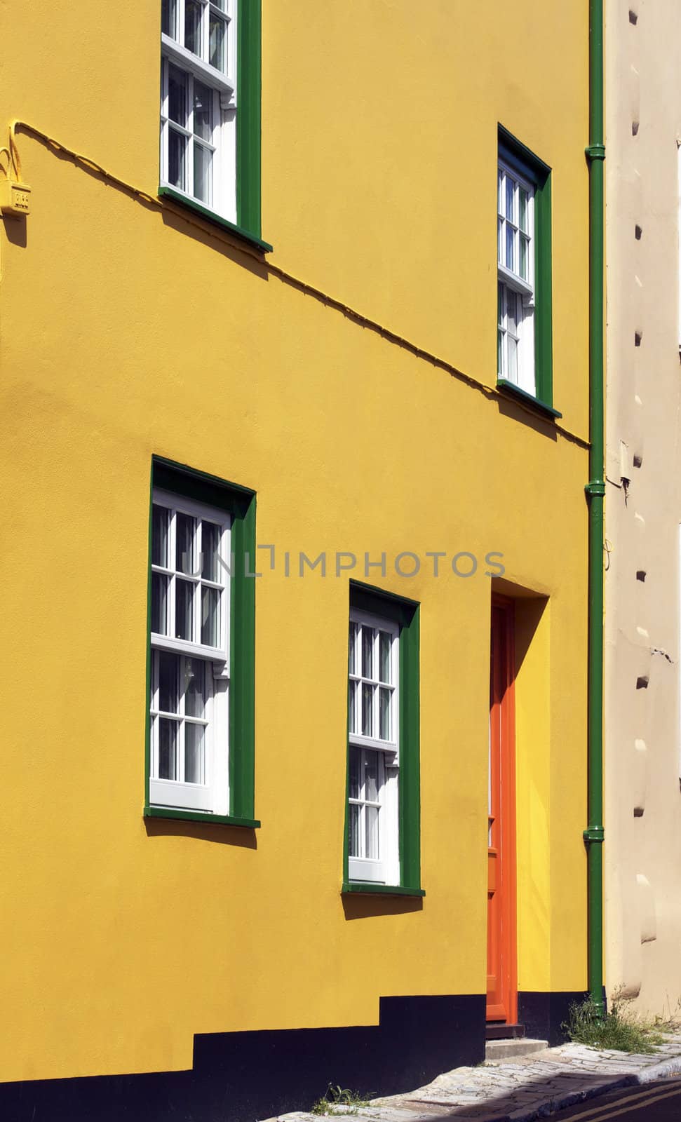 The brightly painted yellow frontage of a coastal cottage. Located in the coastal town of Lyme Regis in Dorset, England. Window frames painted in bright green, with a vivid orange painted door.