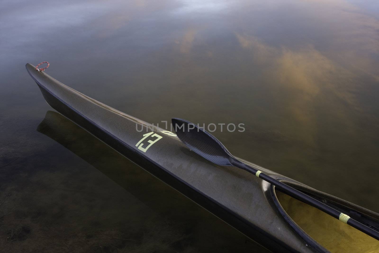 bow of sea racing kayak (lightweight and slim carbon kevlar fiber design) with a wing paddle across cockpit on calm water with cloud reflections, temporary racing number 13 on front deck, wet after paddling

l

eightweight carbon kevlar racing sea kayak with wing paddle on a front deck 