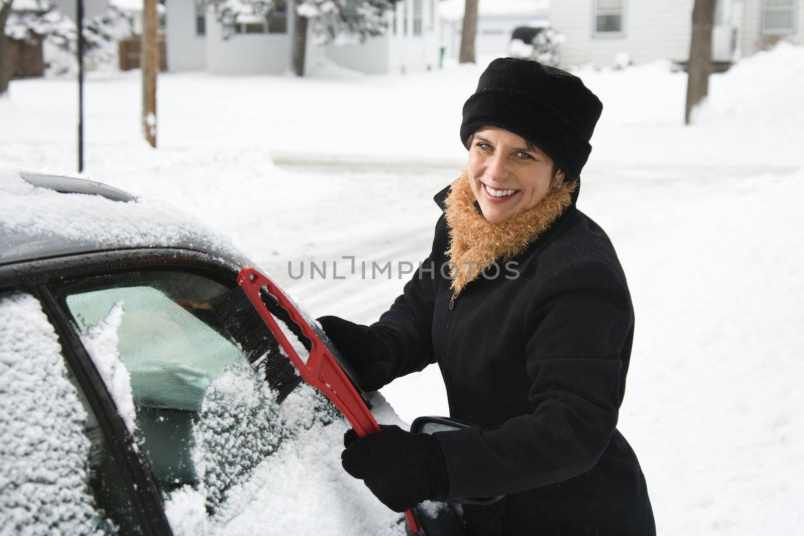Caucasian mid adult woman scraping ice off car windshield and smiling at viewer.