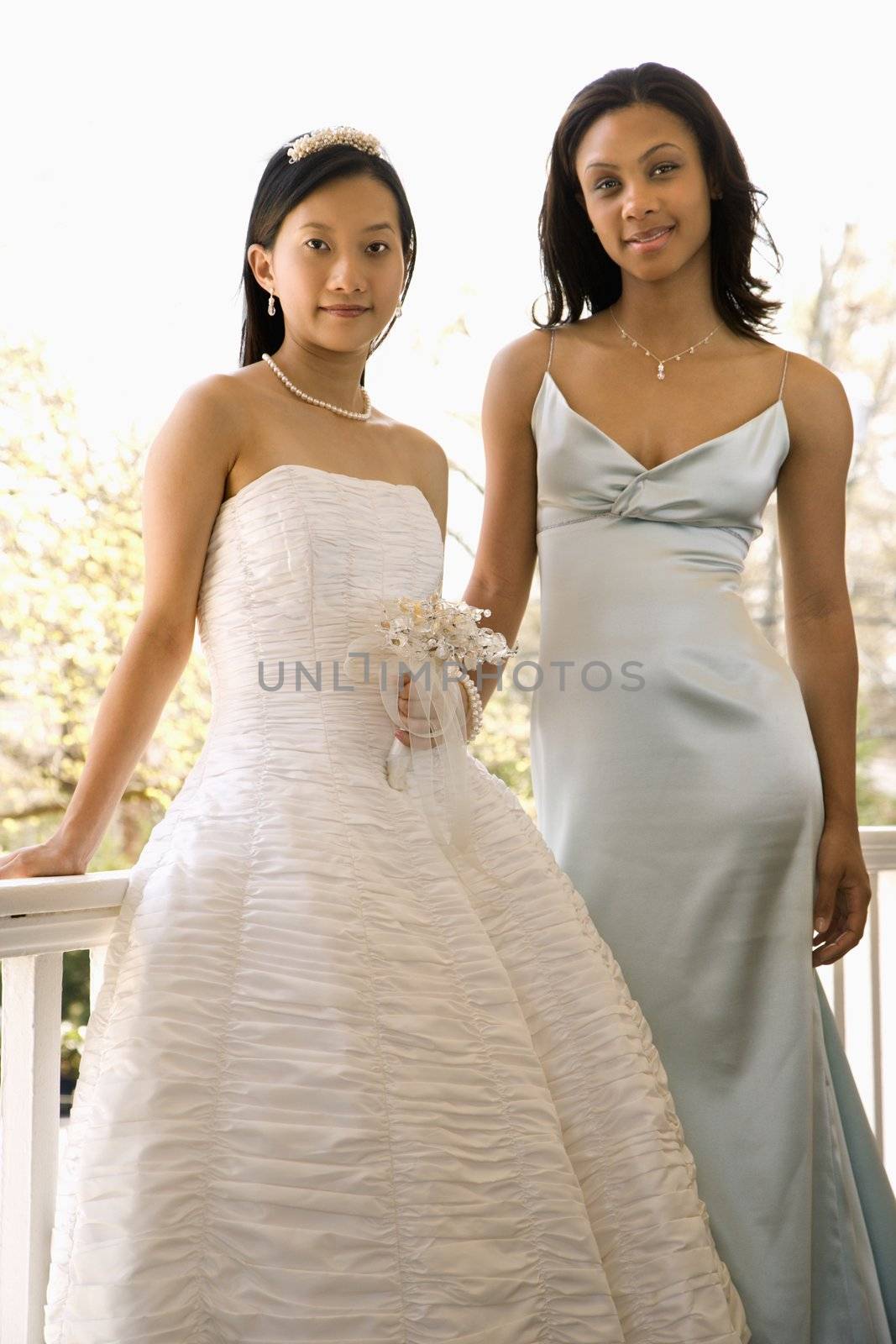 A portrait of a African-American maid of honor and Asian bride leaning against railing.