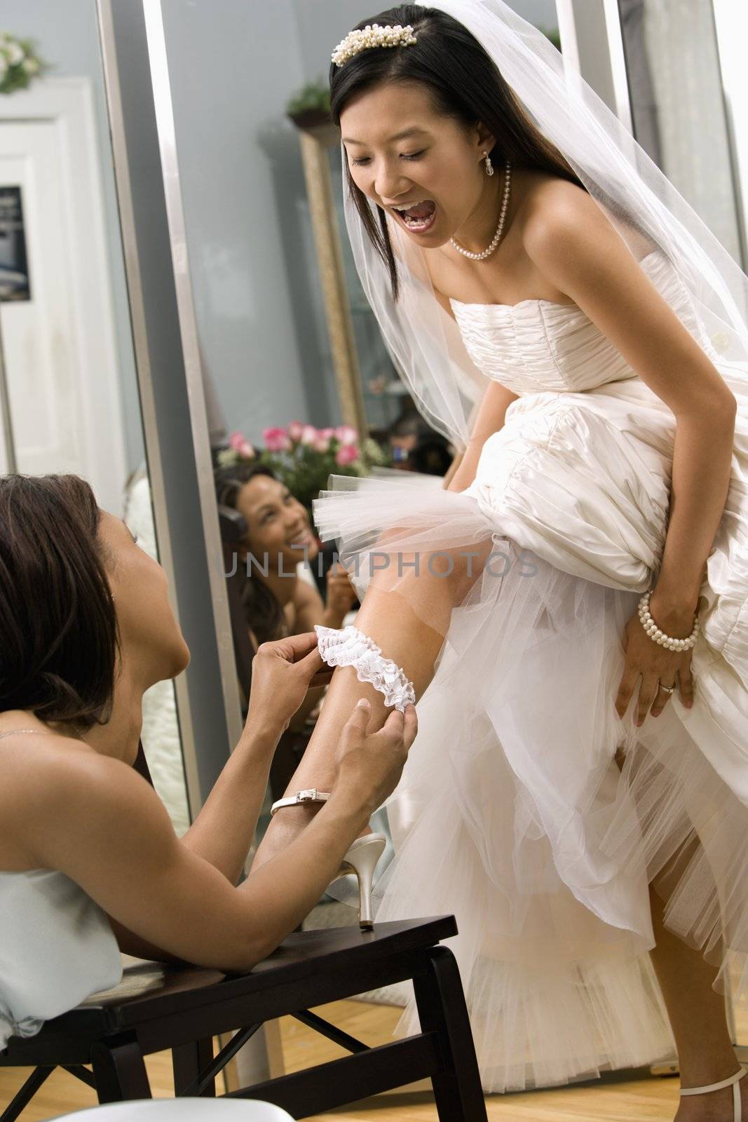 African-American bridesmaid placing garter on Asian bride's leg.