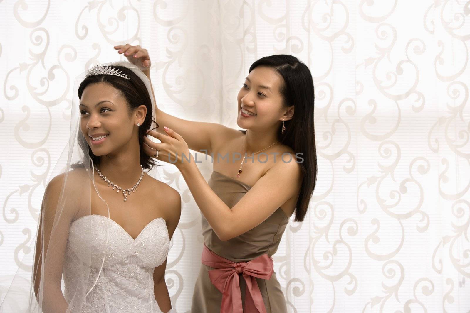 Asian bridesmaid adjusting veil of African-American bride.