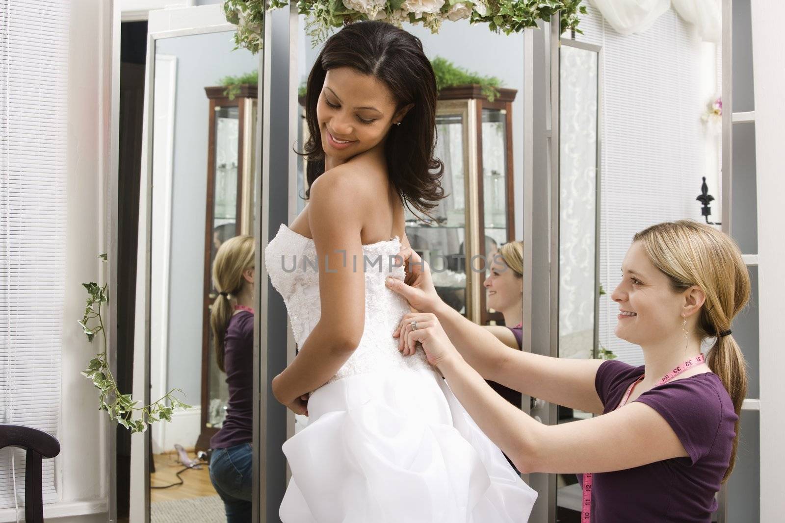 Caucasian seamstress helping African-American bride in bridal shop.