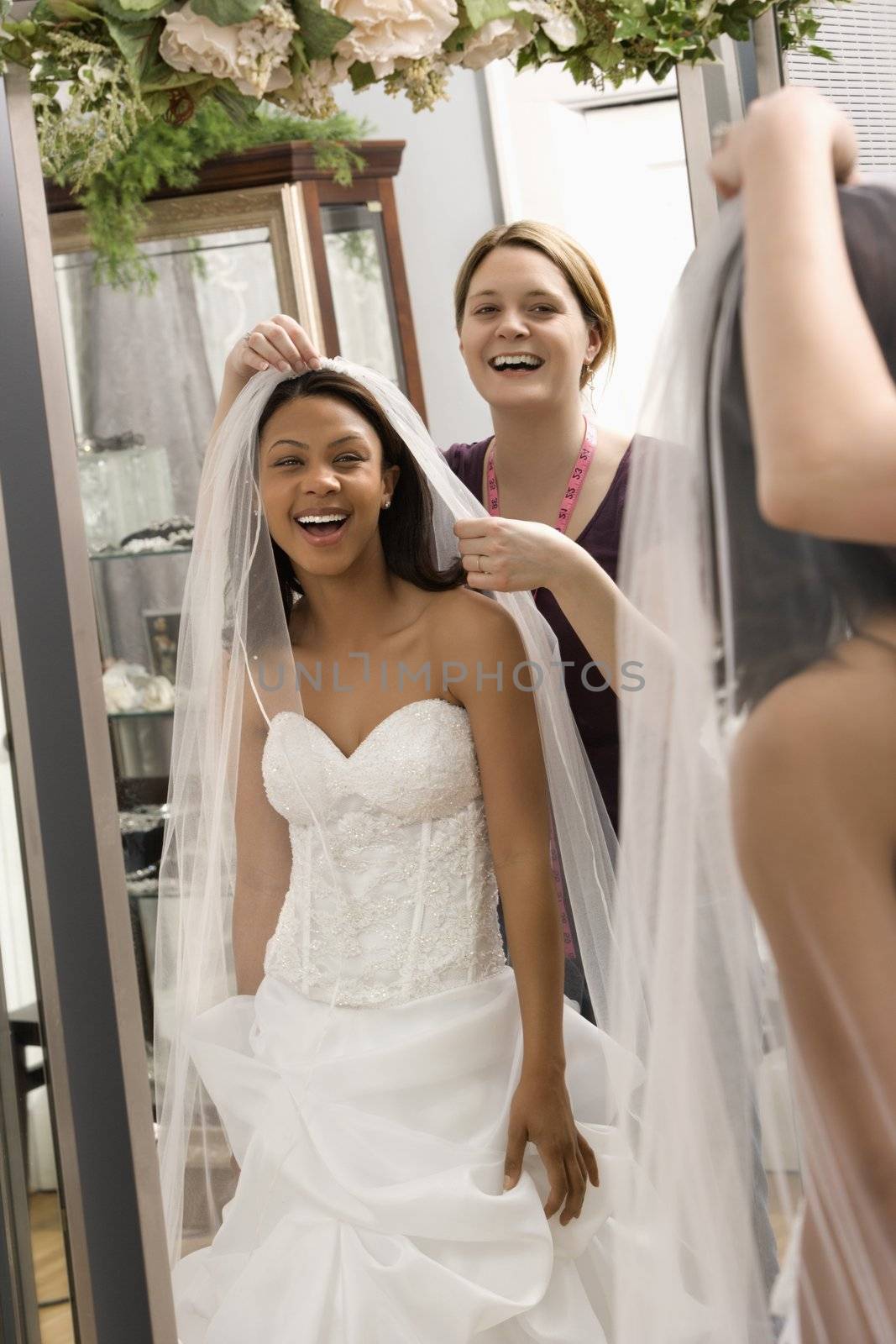 Caucasian seamstress helping African-American bride with veil in bridal shop.