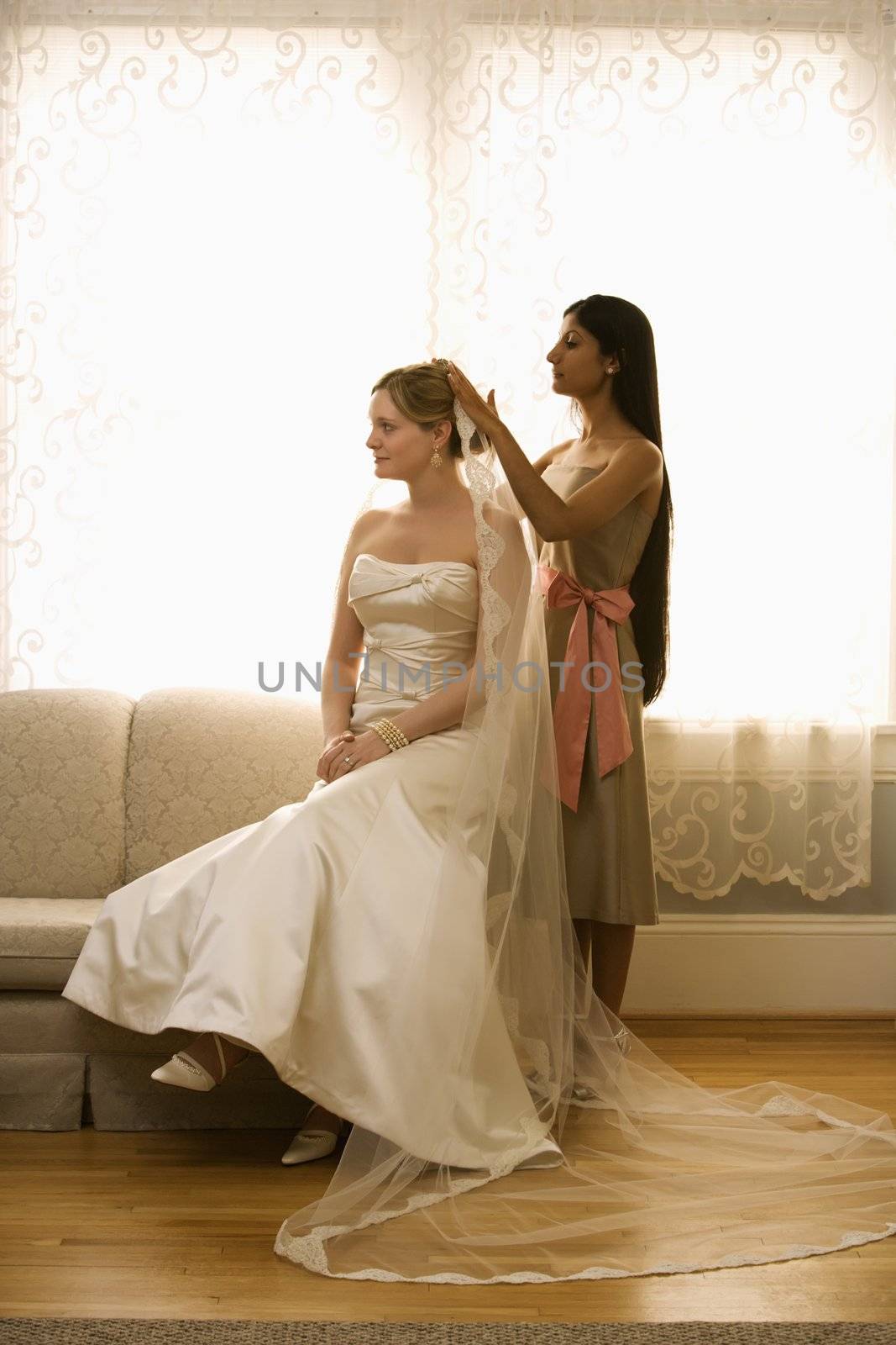 Indian bridesmaid placing veil on Caucasian bride.