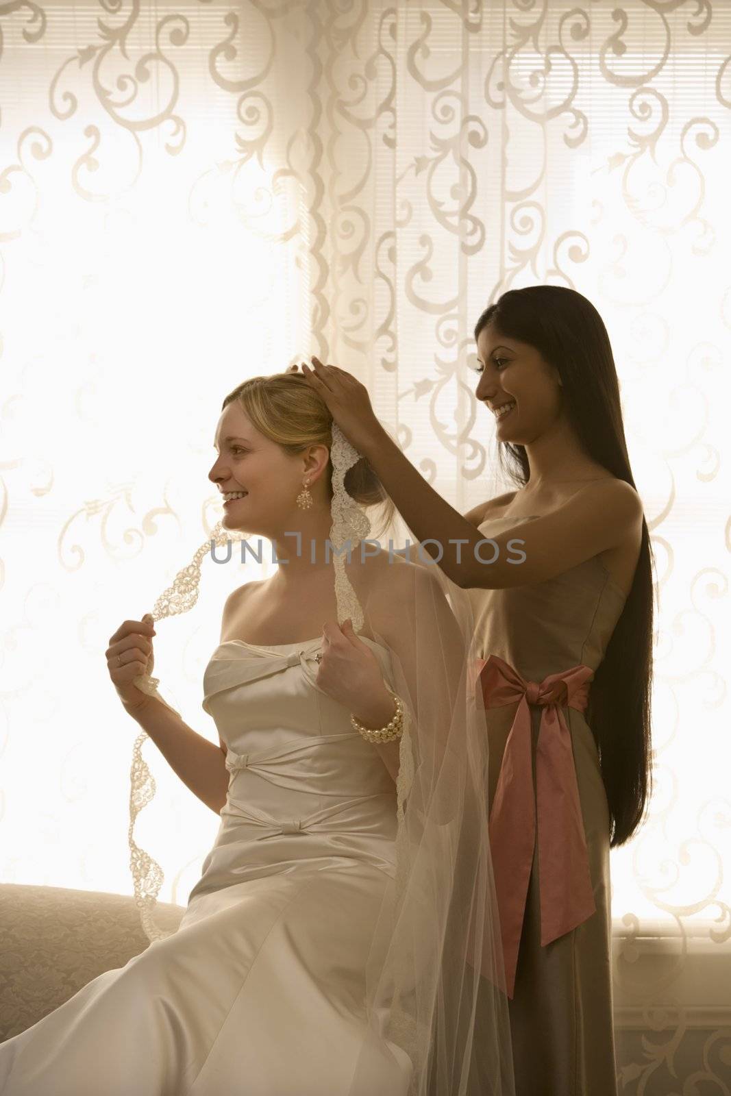 Indian bridesmaid placing veil on Caucasian bride.