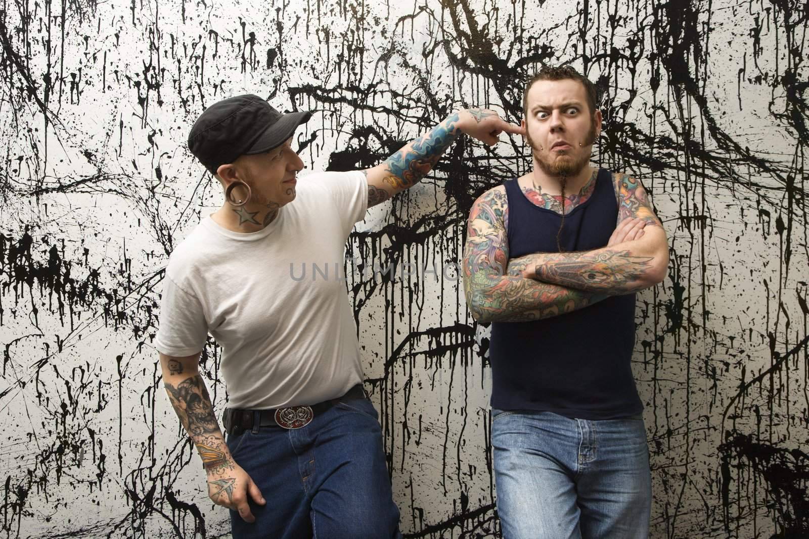 Two Caucasian tattooed men standing against paint splattered background.