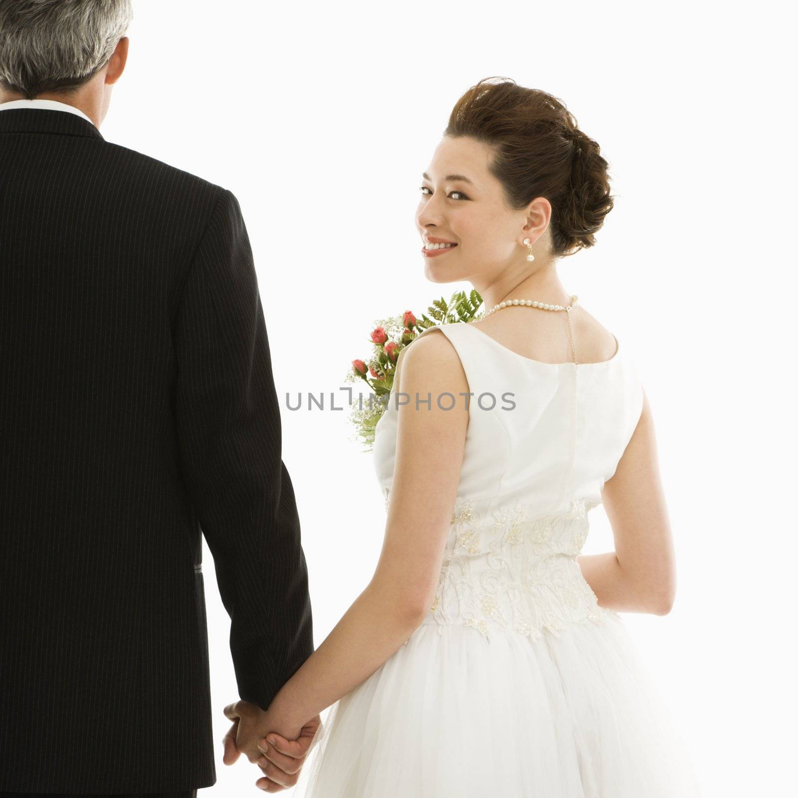 Asian bride looking over her shoulder while holding Caucasian groom's hand.