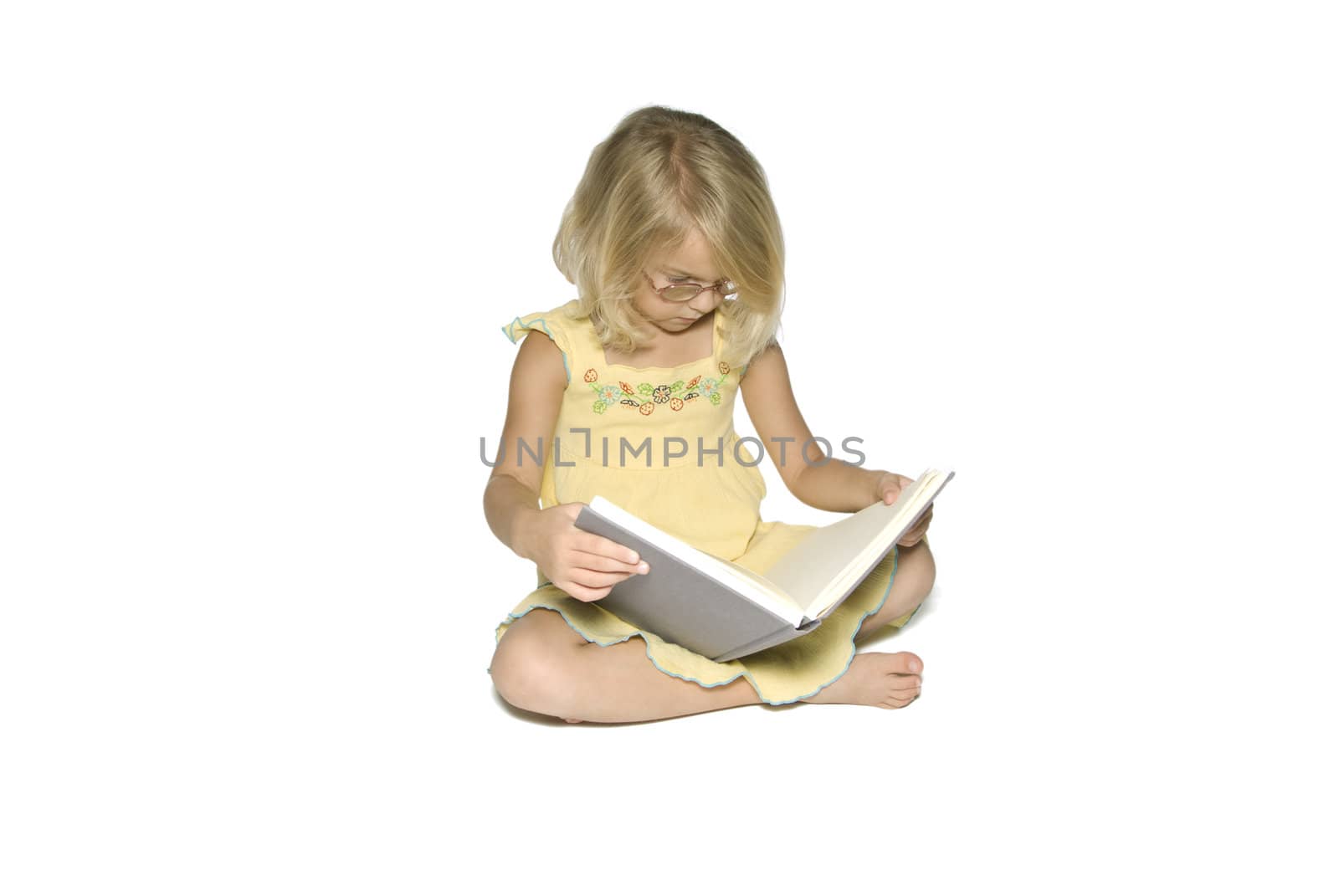 A young blonde girl sitting crosslegged while reading a textbook.  Isolated on a white background