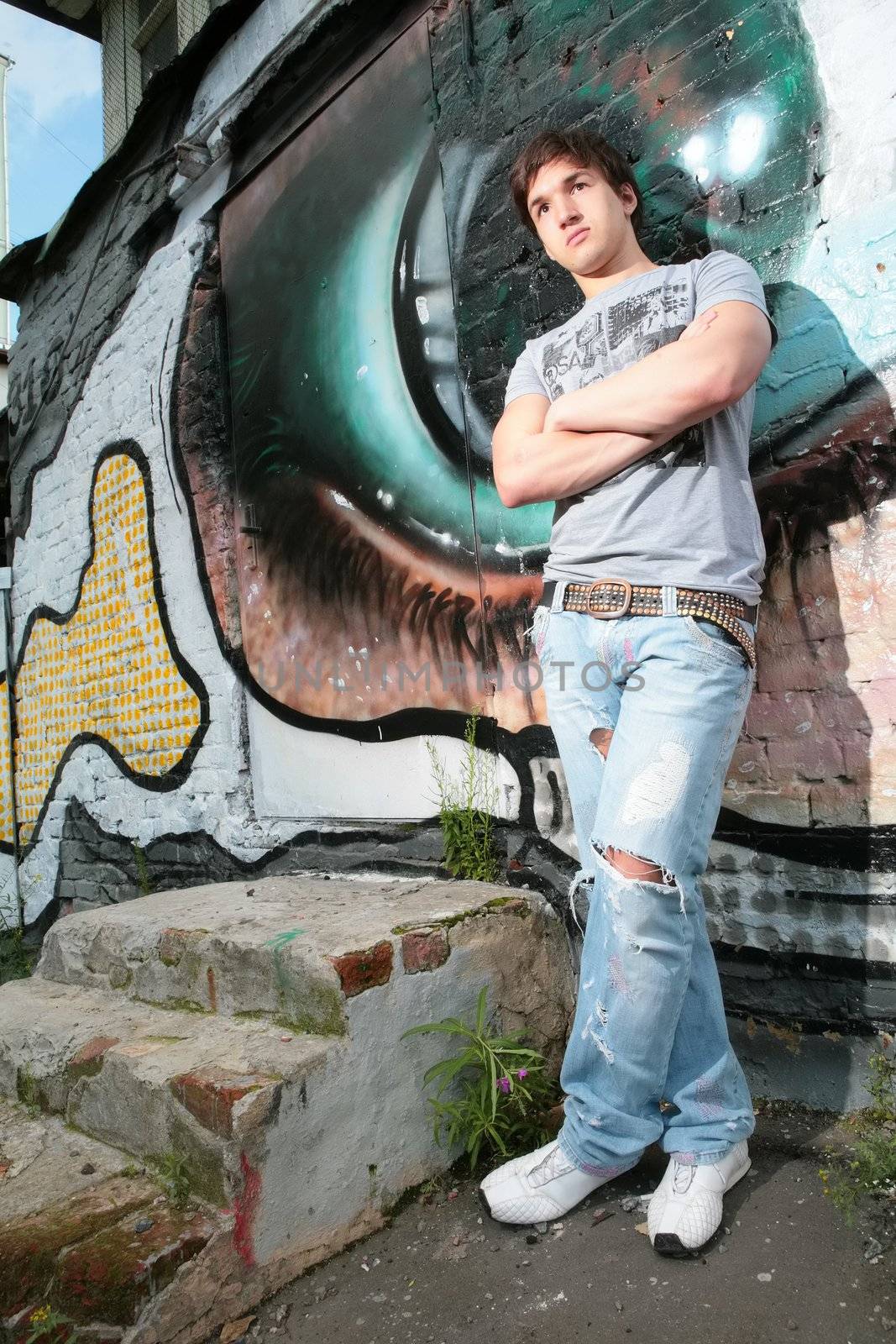 young man stands near graffiti and crossed hands and legs