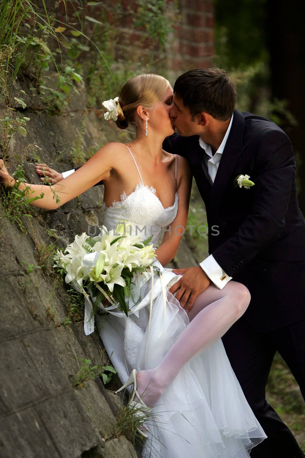 Kissing newlywed couple leaning against wall in countryside.