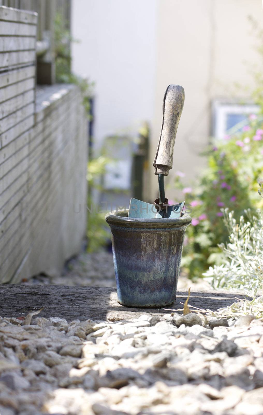 A small hand held trowel with a wooden handle, standing in a glazed blue coloured plant pot. A low angled view looking down a stoney garden path with wooden decking to left of image.