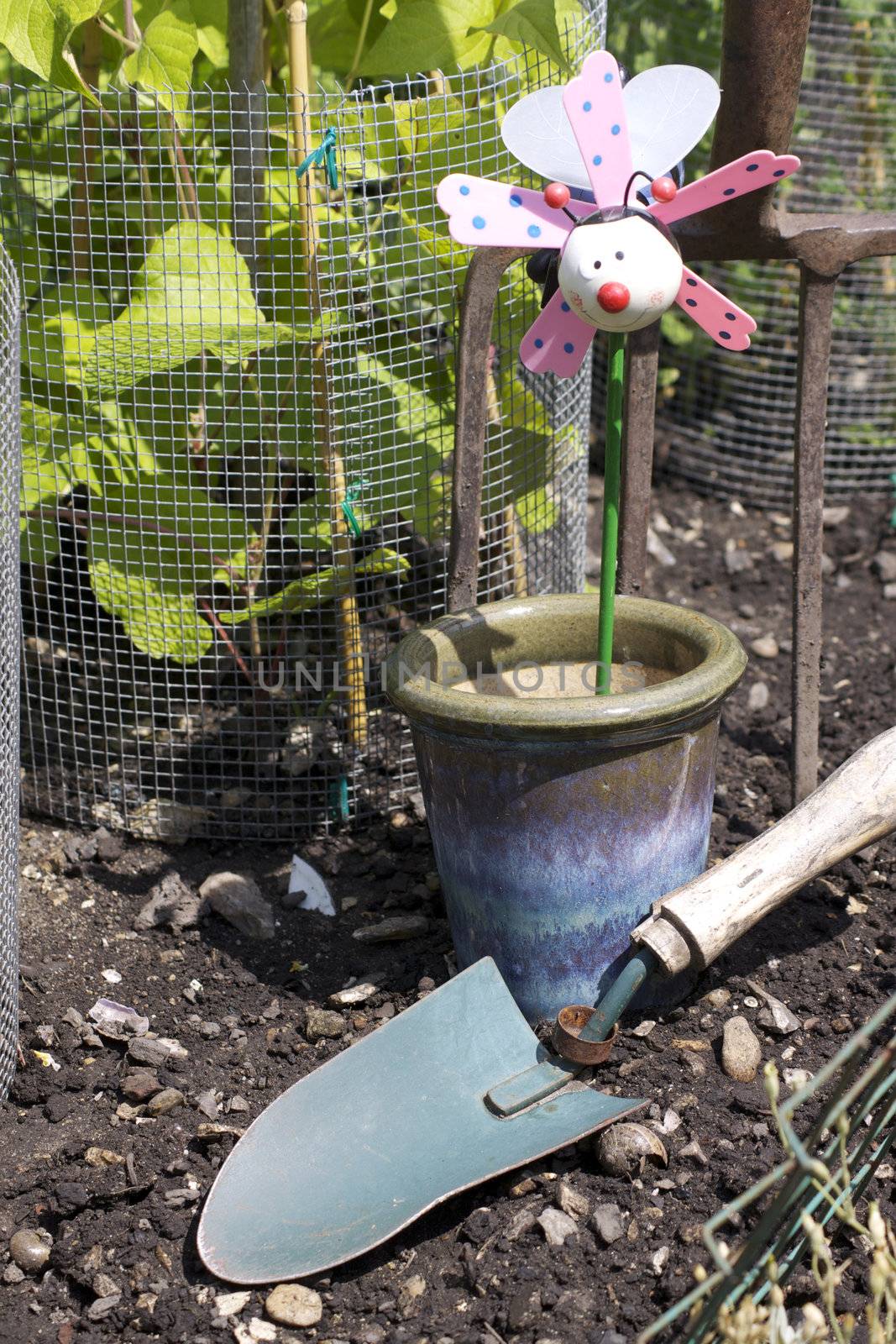 A childs plant pot decoration of a stylised bee. Set in a blue glazed ceramic plant pot in a vegetable garden amongst growing runner beans. A small hand held garden trowel lies in front of the pot.