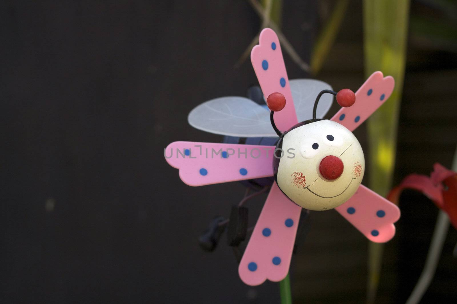 A childs plastic plant pot toy decoration of a stylised bee with pink wings. Set in a garden flower pot.