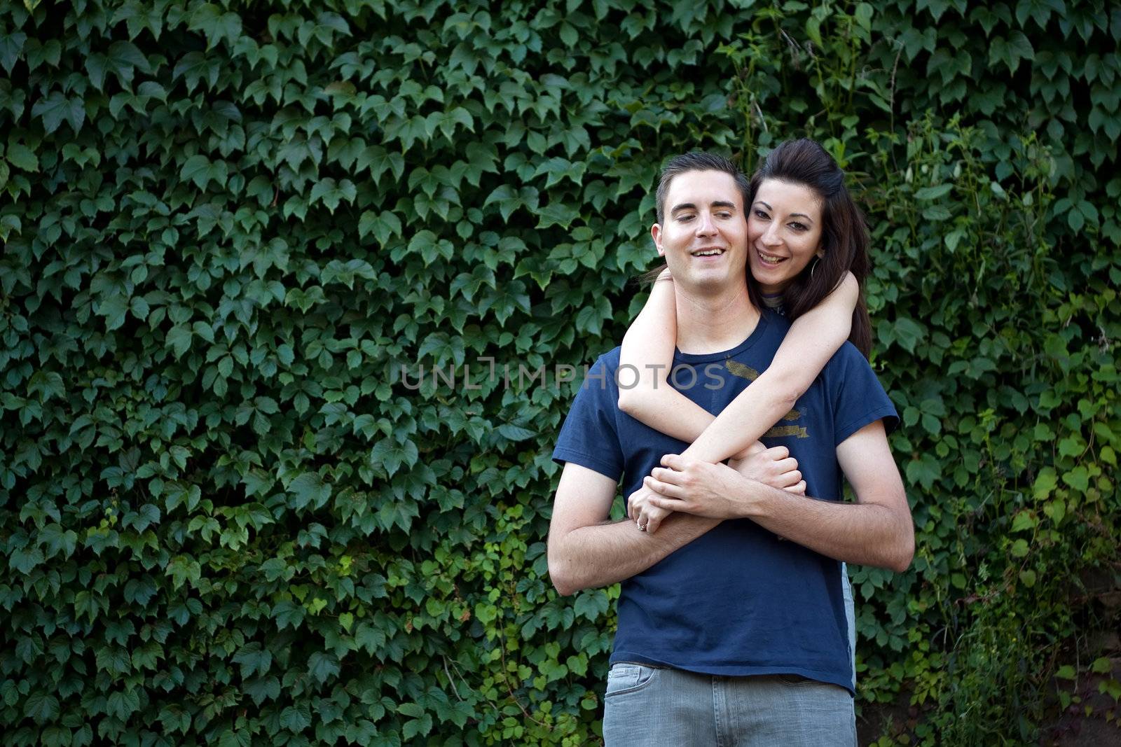A happy young couple in their mid 20s together outdoors in front of some green ivy.