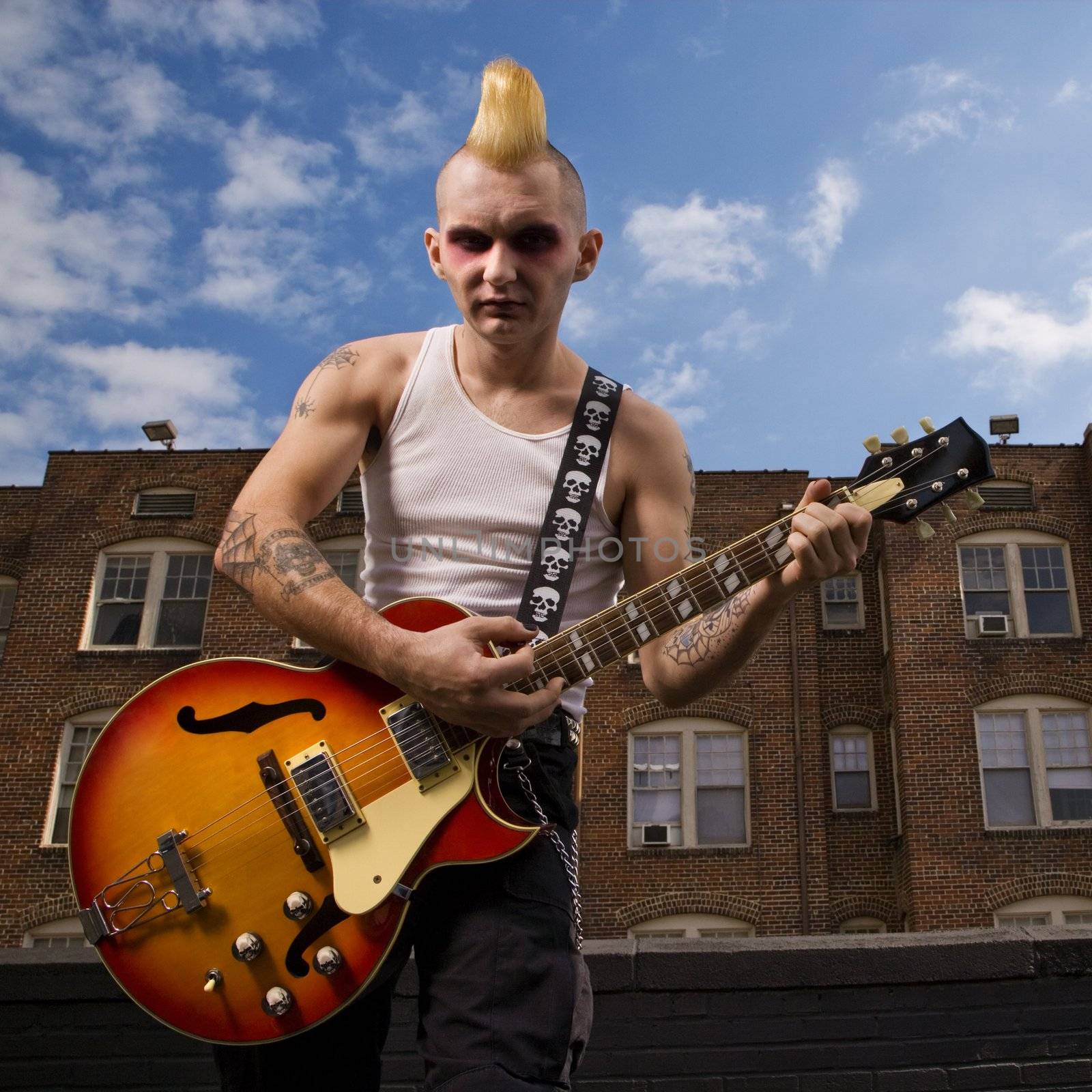 Portrait of a mid-adult Caucasian male punk playing guitar with building in background.