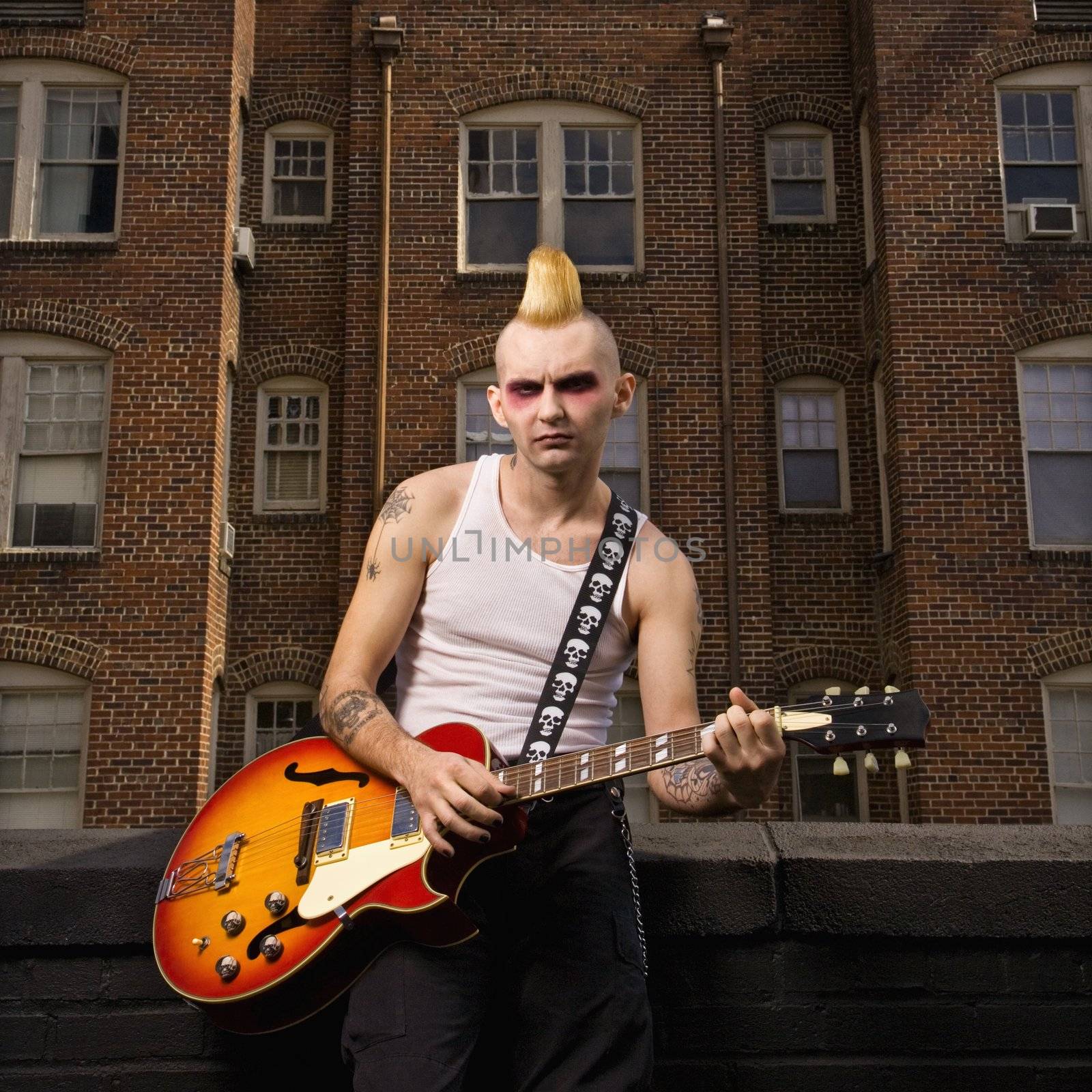 Portrait of a mid-adult Caucasian male punk playing guitar with building in background.