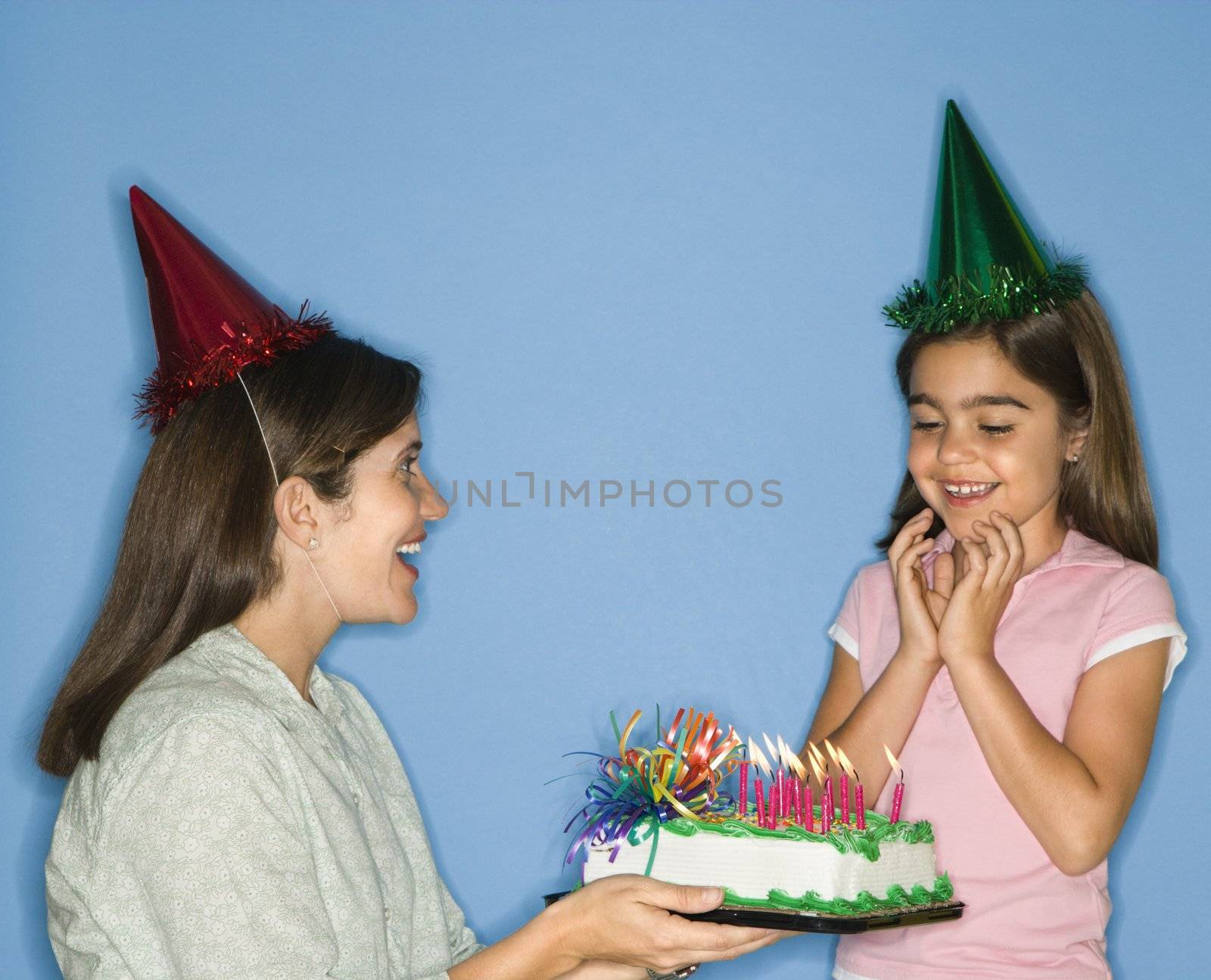 Girl wearing party hat with mother holding birthday cake.