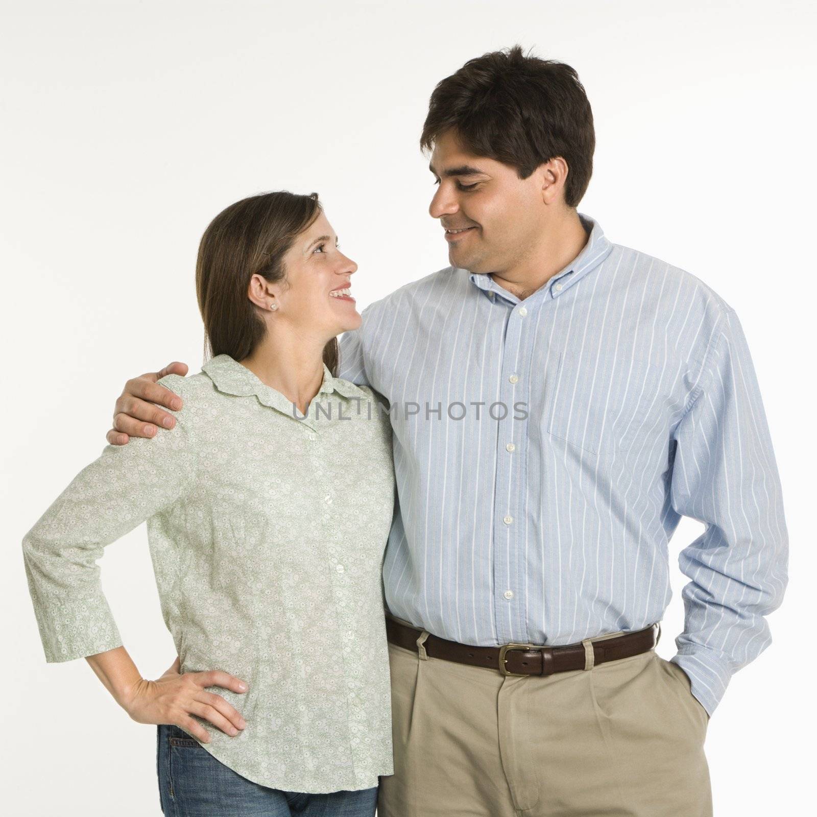 Couple standing smiling at eachother against white background.