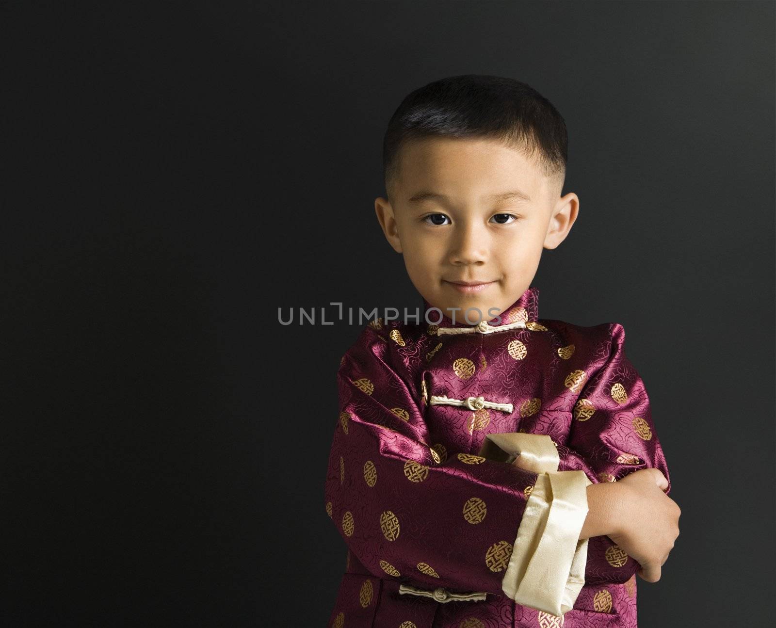 Asian boy in traditional attire standing against black background.