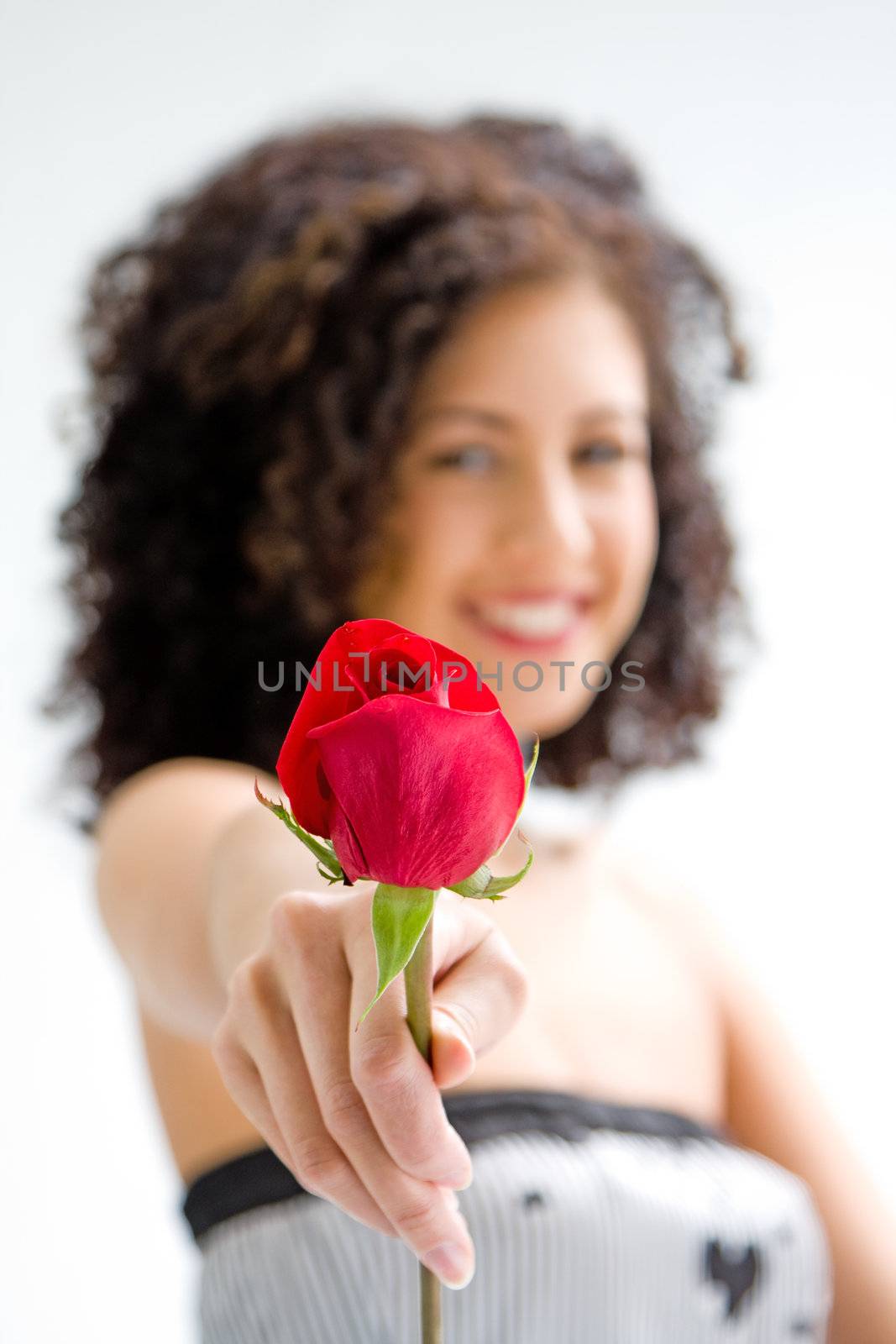 Sincere beautiful young woman with brown curly wild hair and bare shoulders presenting red rose, isolated