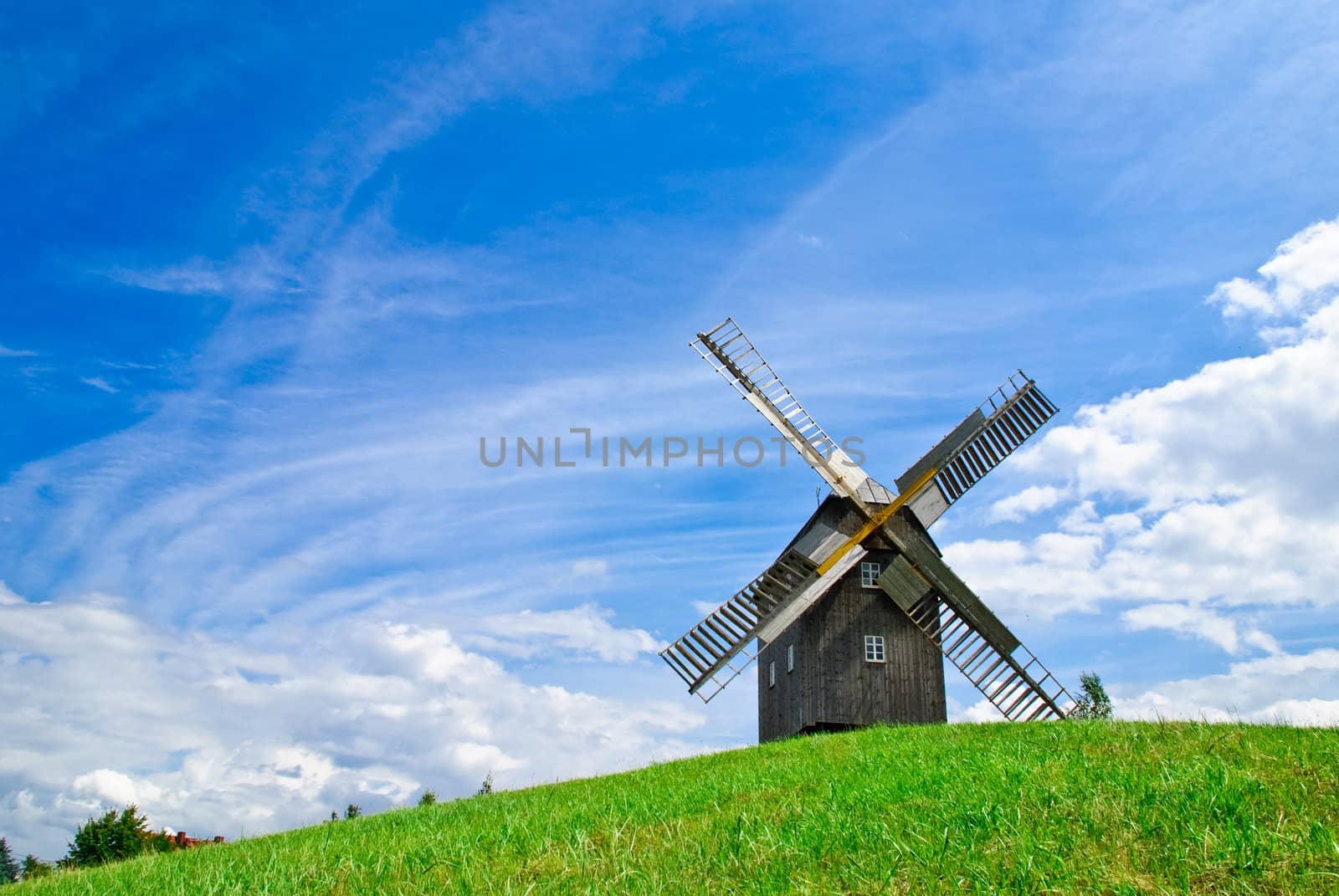 Wooden windmill against the summer blue sky with white clouds on a green hill with a birch