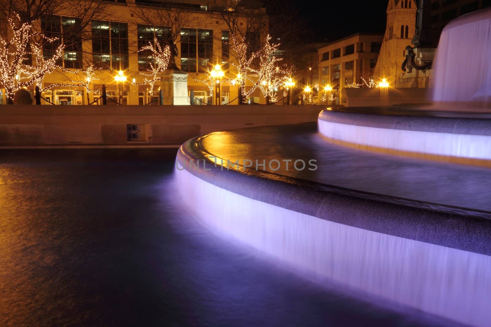 A city fountain eliminated at night with trees covered in lights.