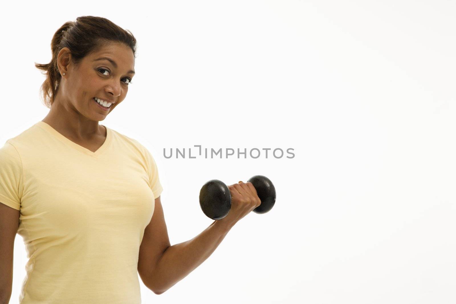 Mid adult multiethnic woman wearing yellow exercise shirt doing arm curls looking at viewer and smiling.