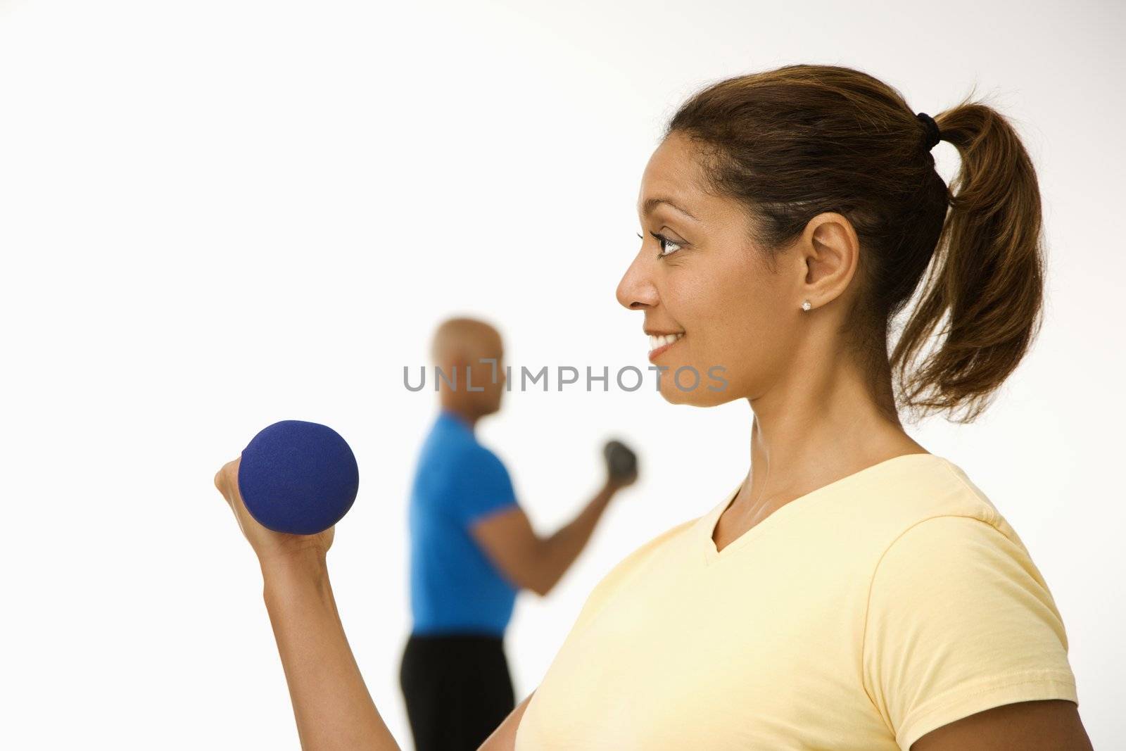 Close up of smiling mid adult multiethnic woman exercising using dumbbells with mid adult multiethnic man in background.