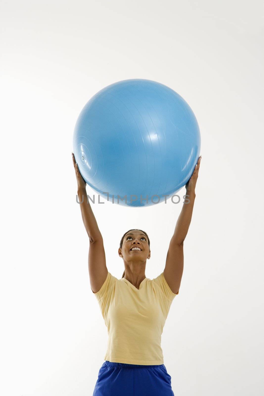 Mid adult multiethnic woman standing and holding blue exercise ball over her head smiling.