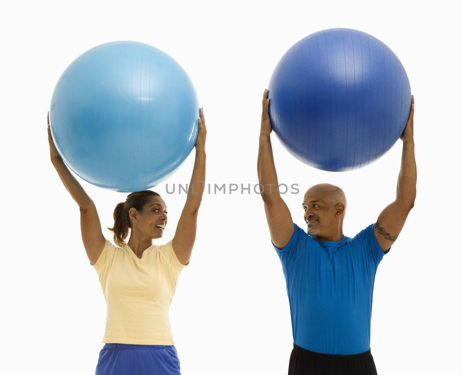 Happy smiling mid adult multiethnic man and woman holding blue exercise balls over their heads looking at each other.