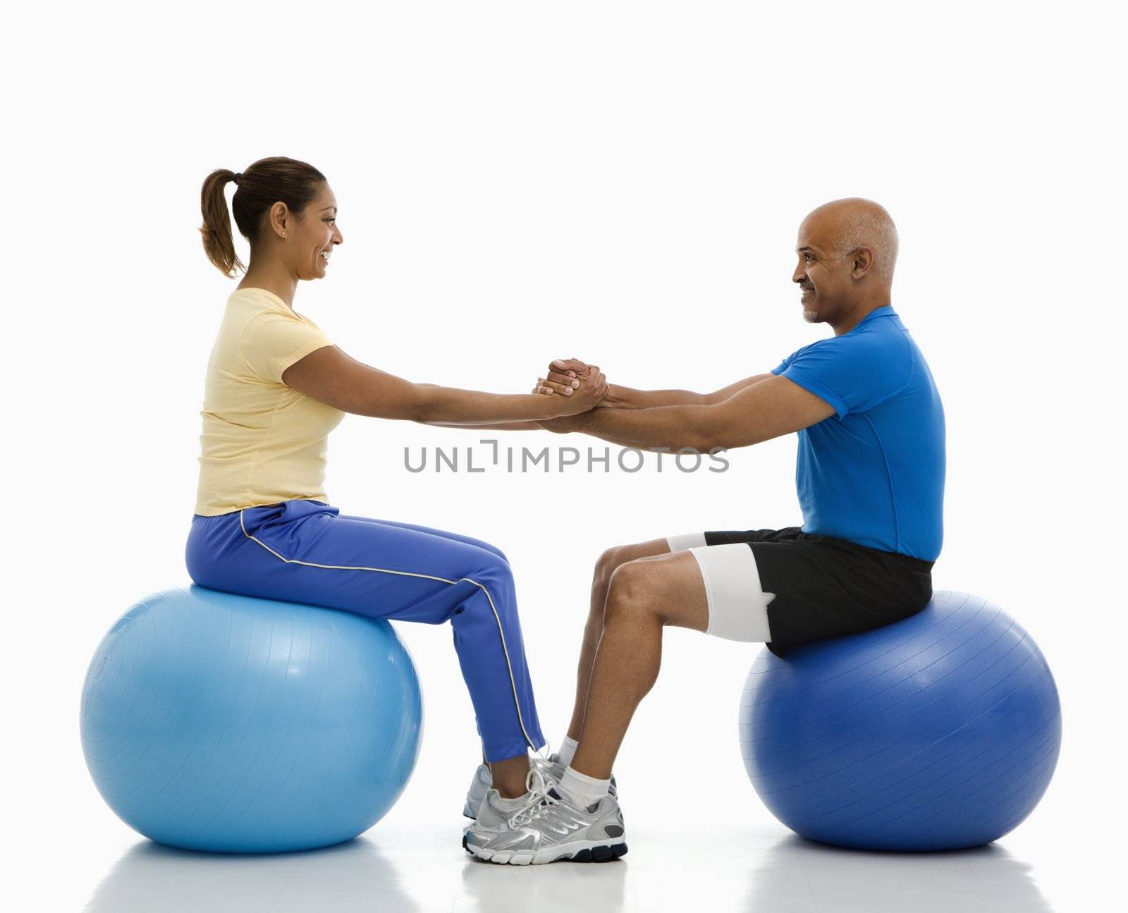 Mid adult multiethnic man and woman balancing on blue exercise balls facing each other with hands and feet locked together.